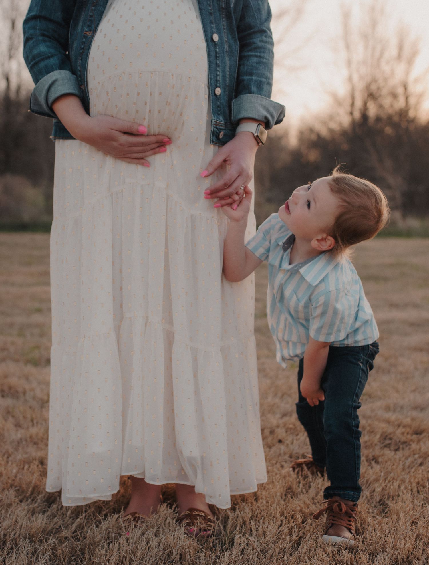 Pregnant mother in white dress holding toddler son's hand in golden field.