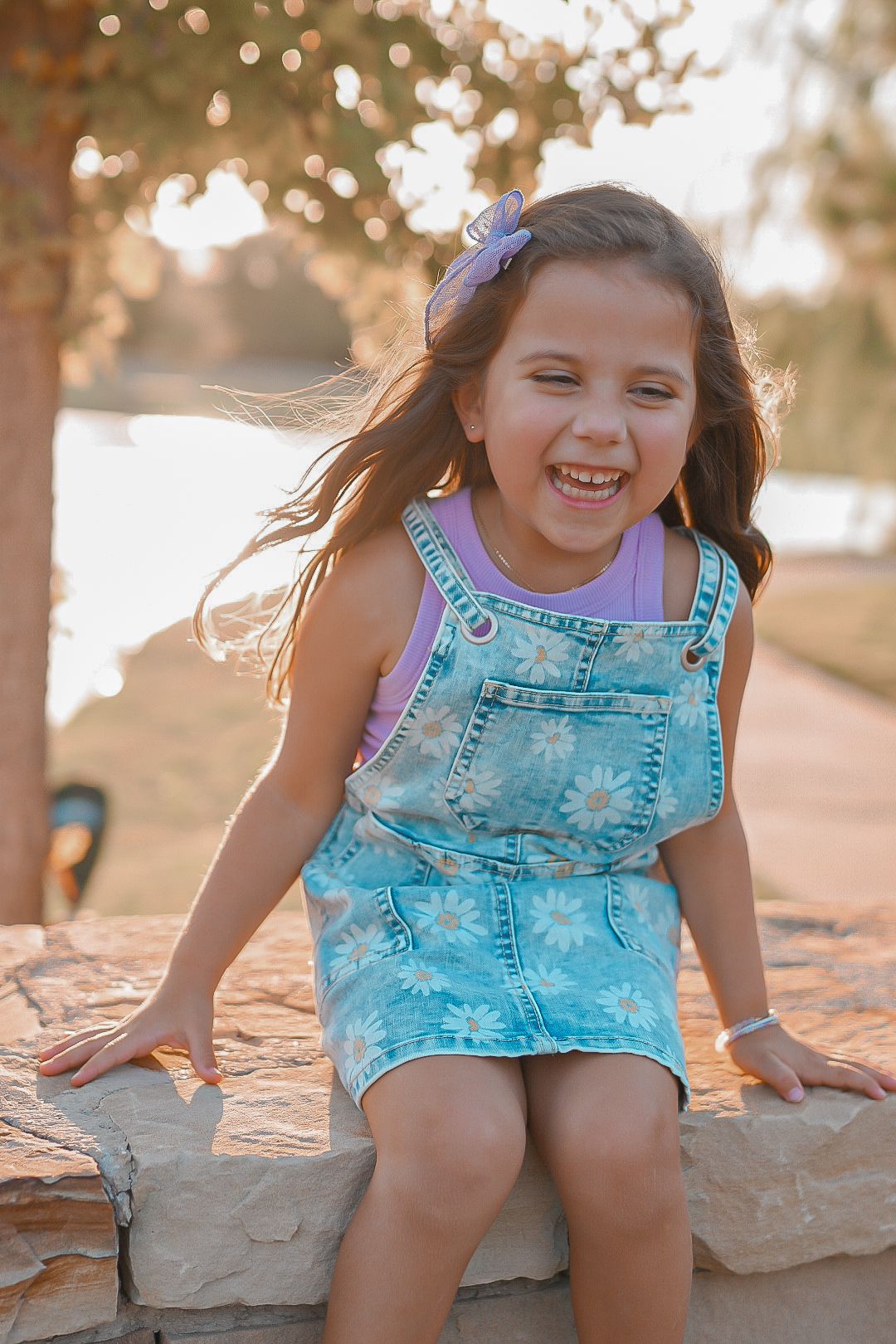 Laughing young girl in floral denim overalls sitting on a stone wall outdoors in golden sunlight.