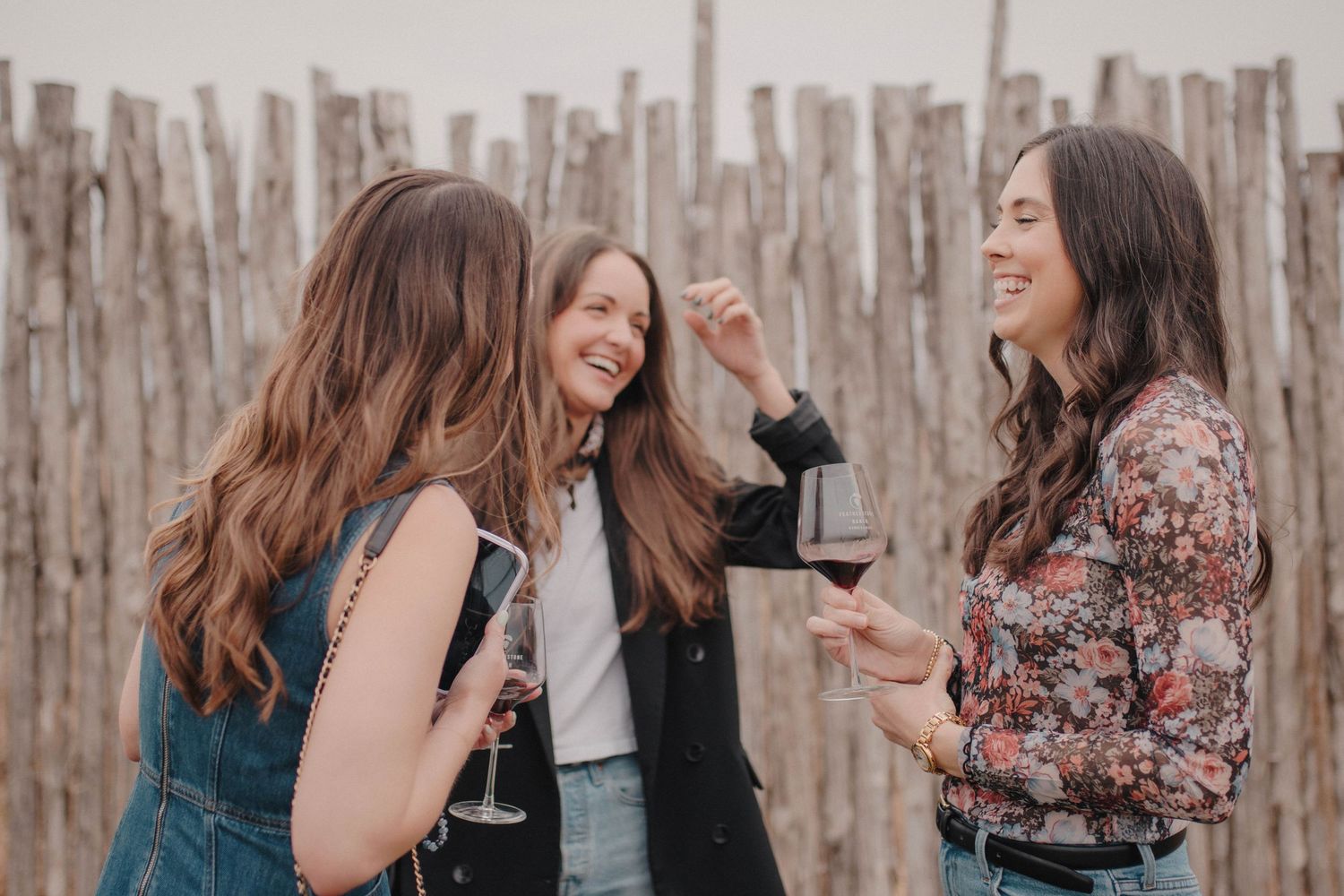 Three women laughing and enjoying wine together outdoors near a rustic wooden fence.