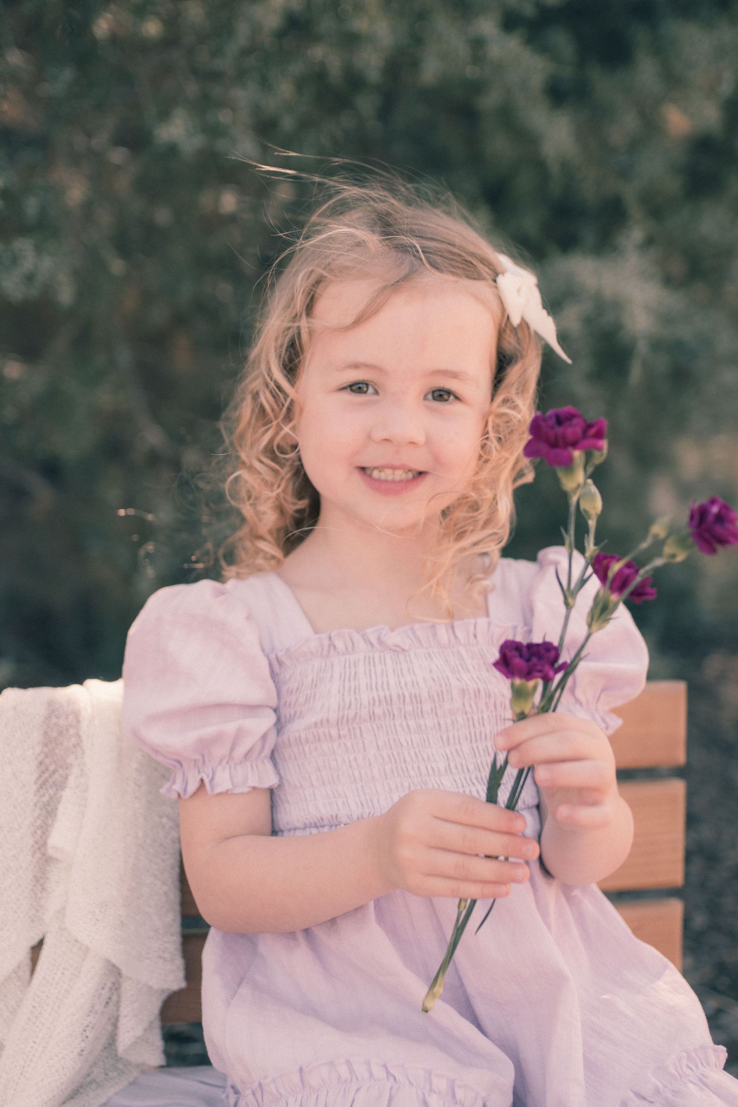 Smiling little girl with curly blonde hair holding purple carnations, wearing a pink smocked dress outdoors.
