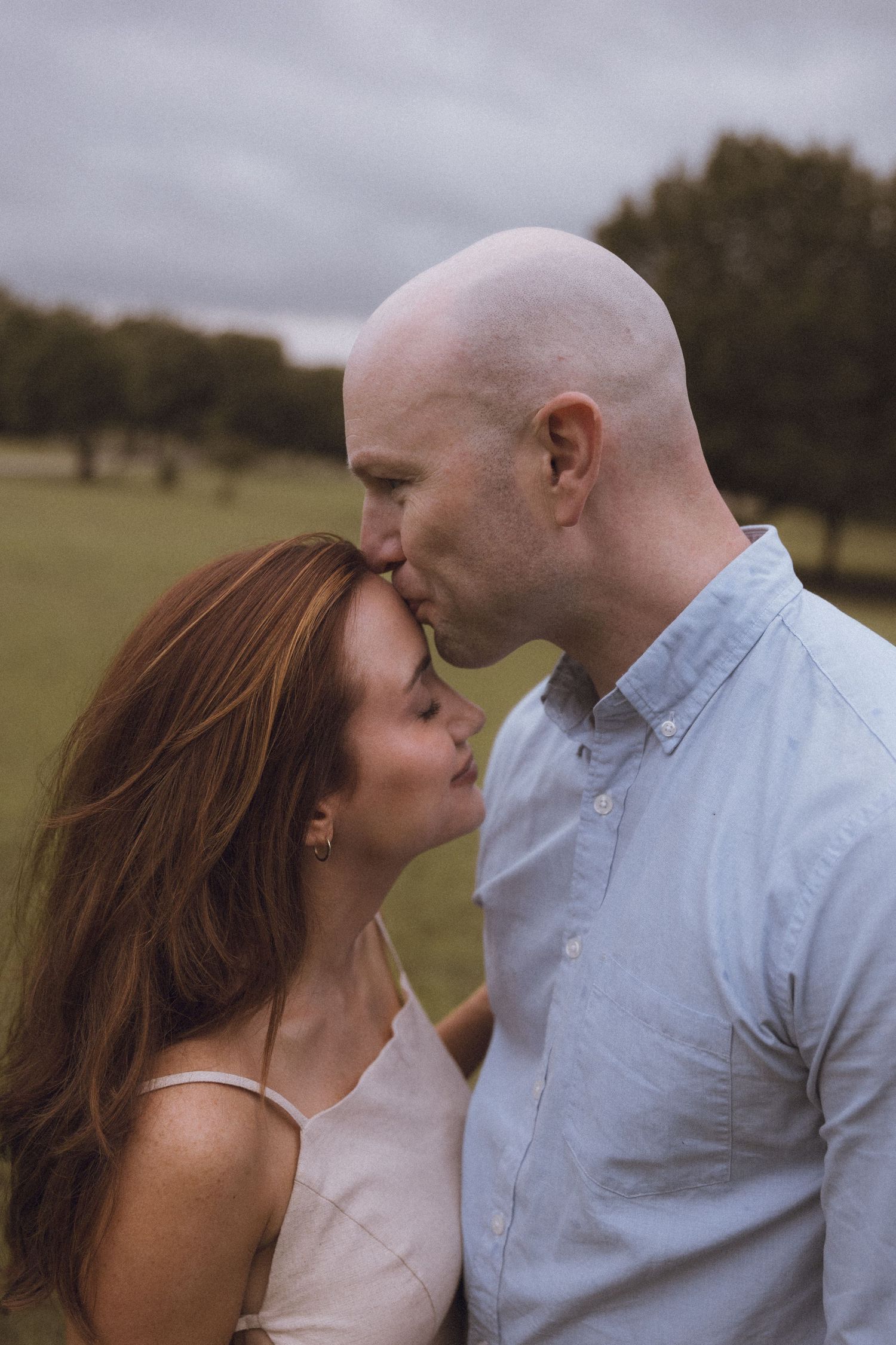 A bald man tenderly kisses a redheaded woman on the forehead in a lush green park under cloudy skies.