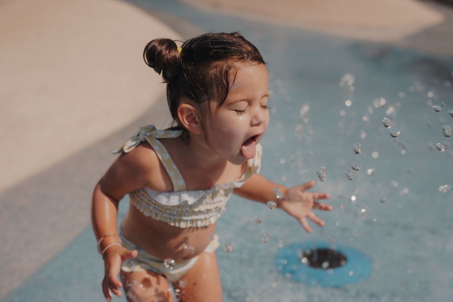 Excited toddler girl in swimsuit playing in splash pad water feature.