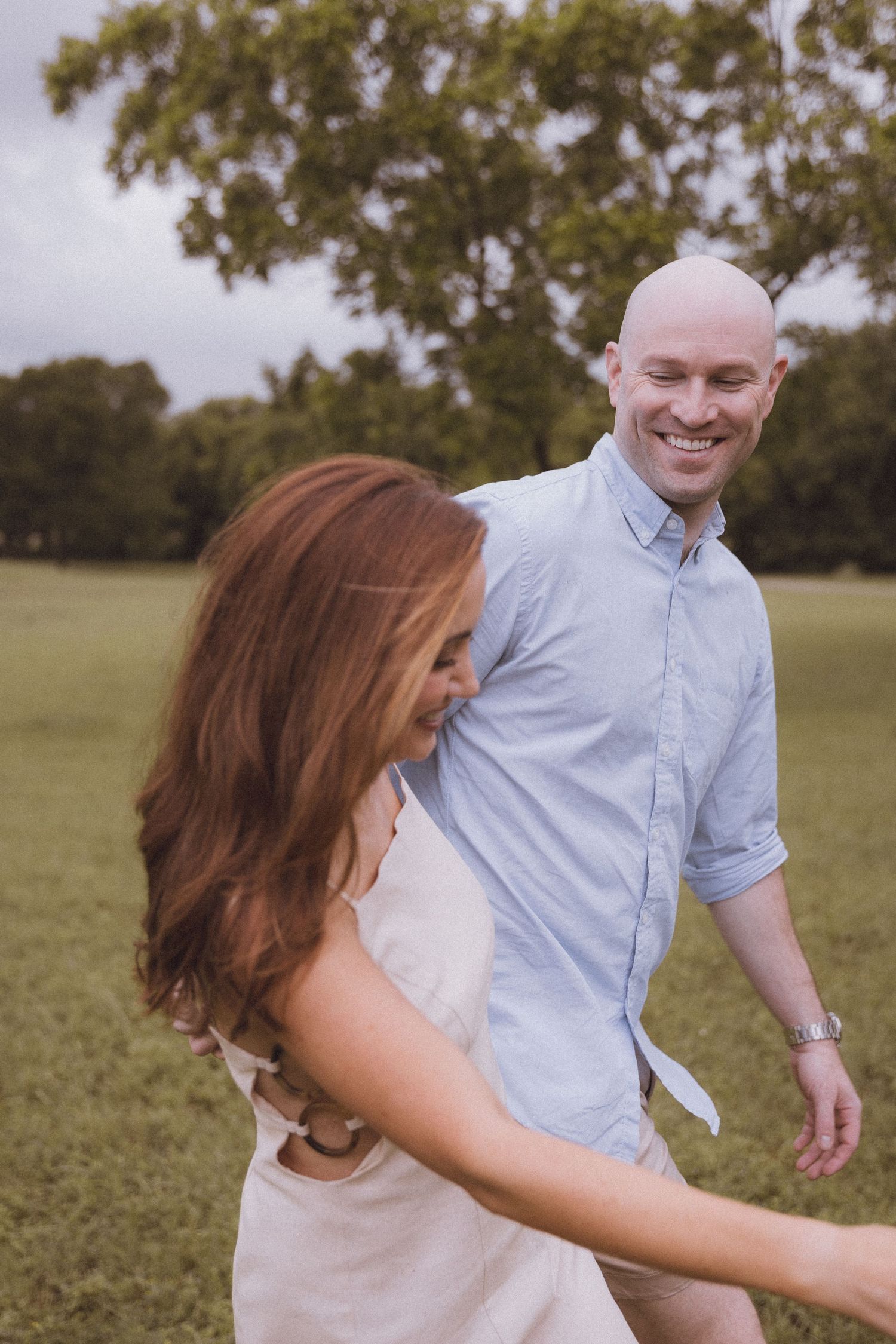 A smiling bald man in a blue shirt dances with a redhead woman in a white dress on a green field.