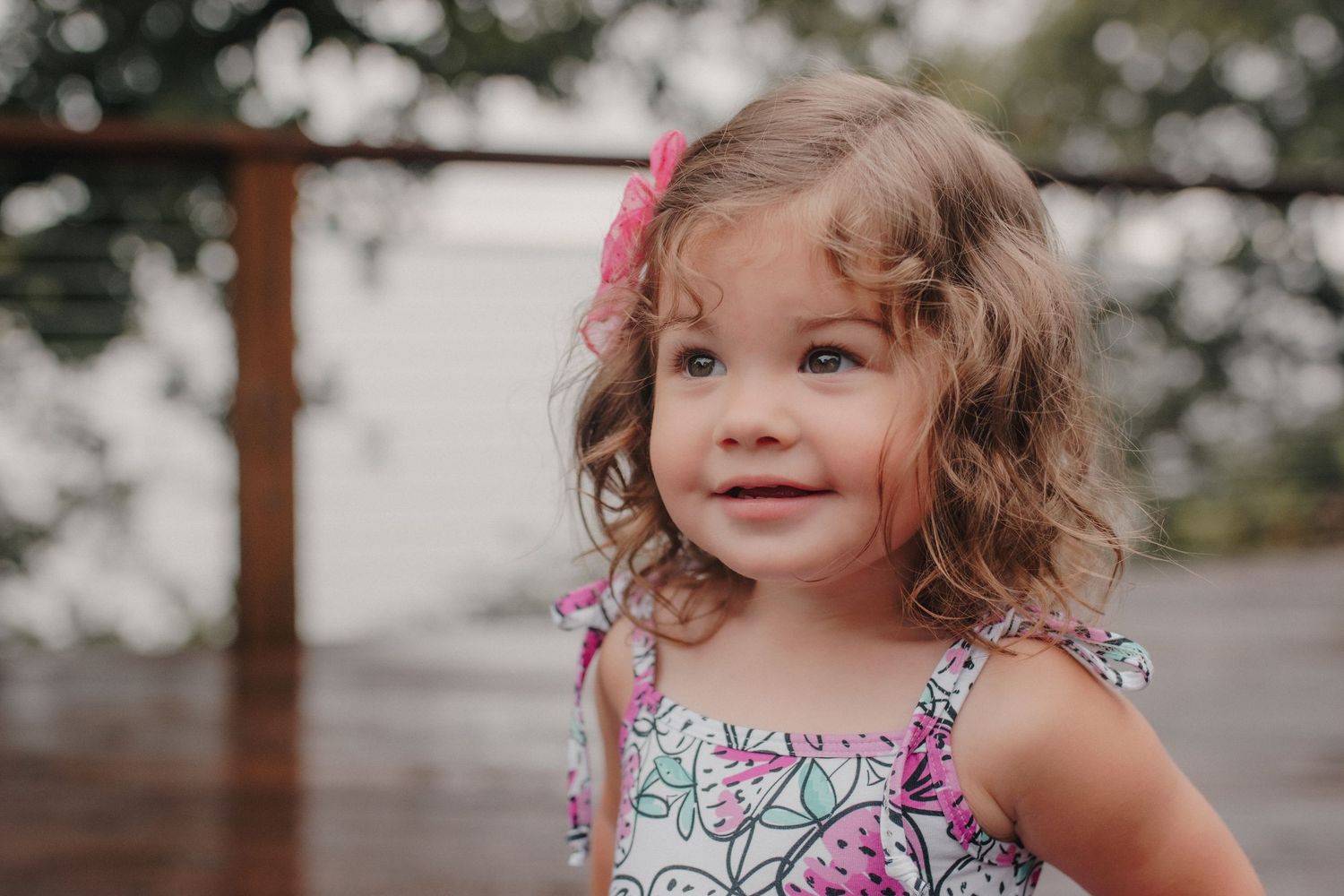 Adorable curly-haired little girl with pink bow smiling at camera outdoors.