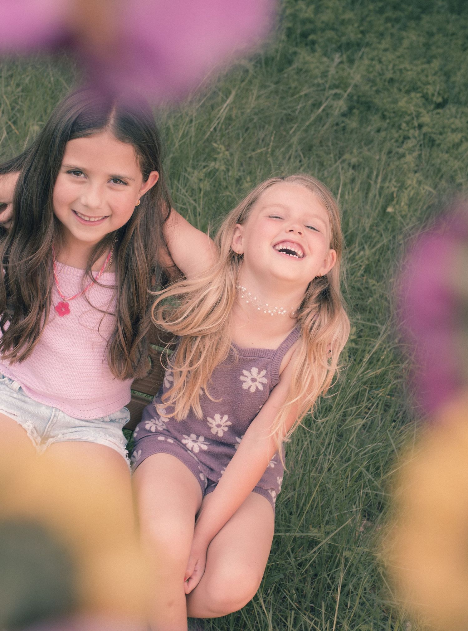 Two happy young girls laughing and sitting together in a flower field on a sunny day.