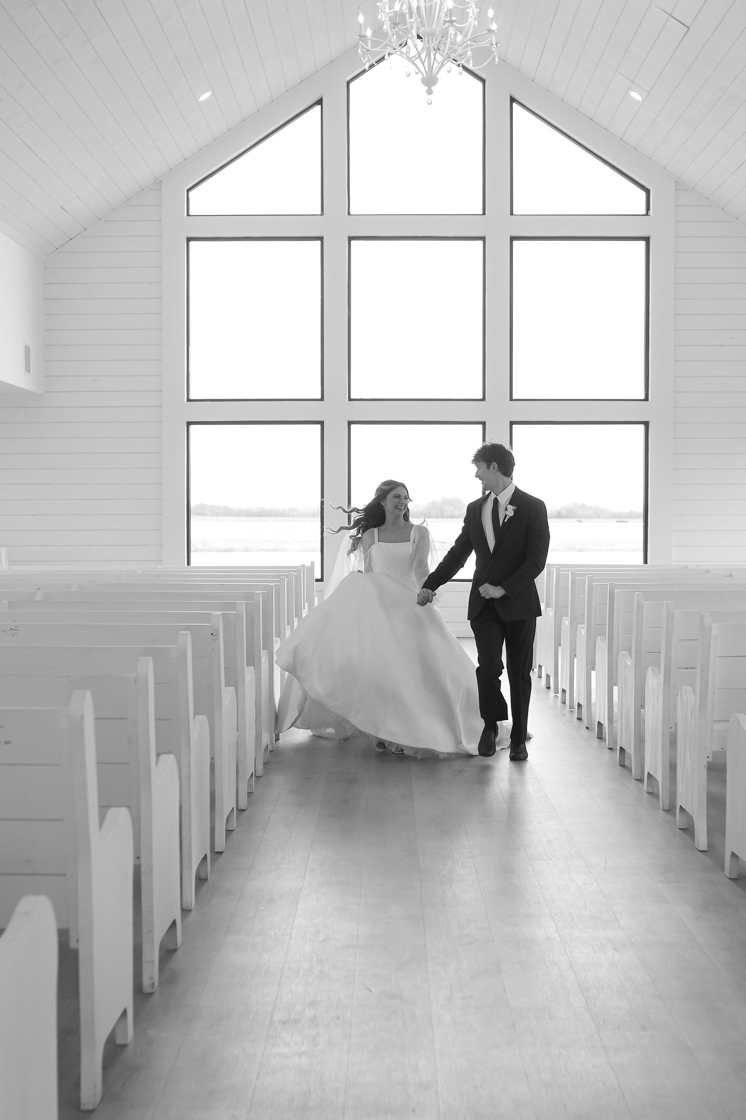 Black and white photo of bride and groom walking down chapel aisle with large windows and chandelier overhead.