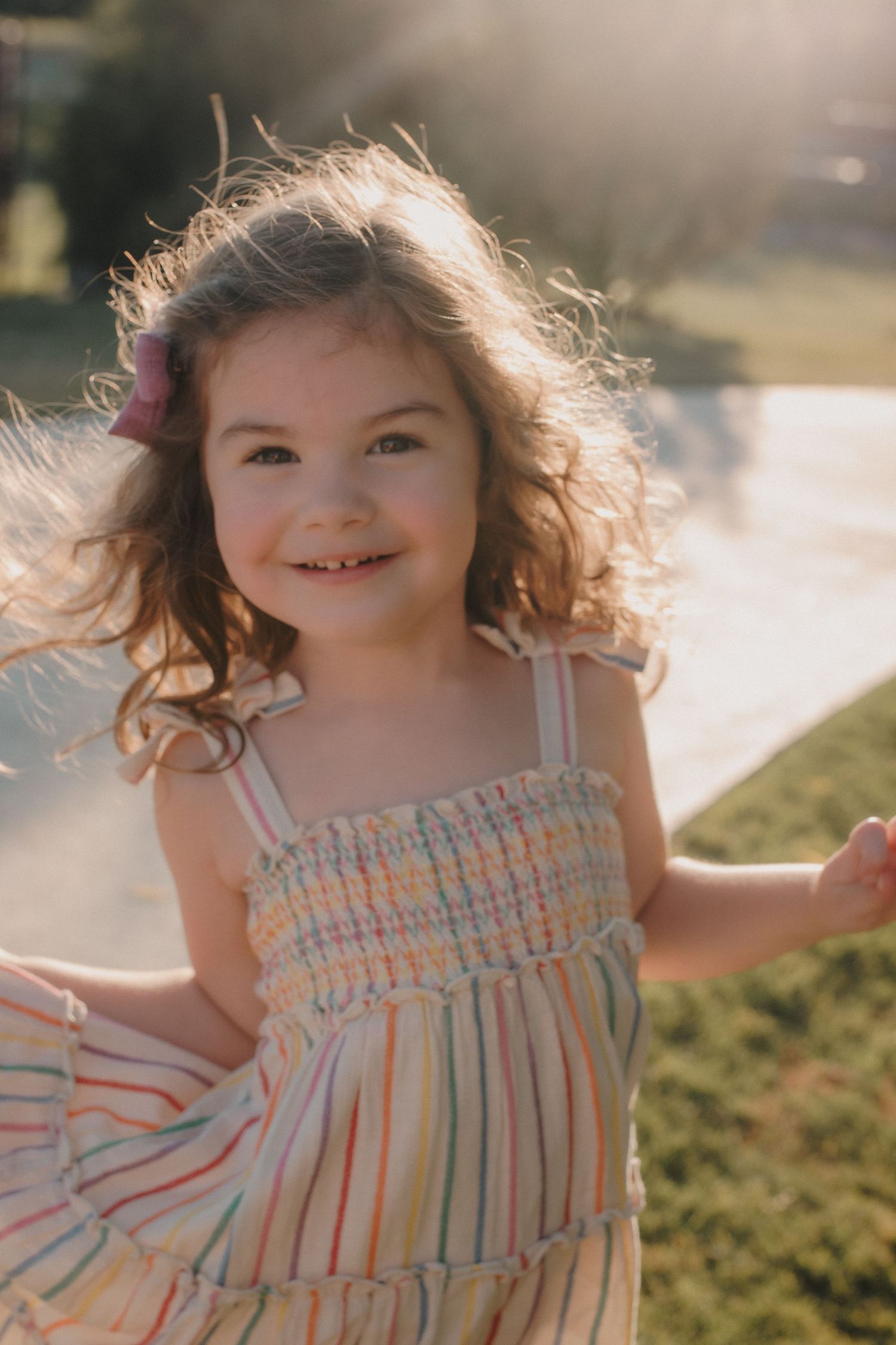 Joyful little girl in colorful striped smocked dress twirling in golden afternoon sunlight.