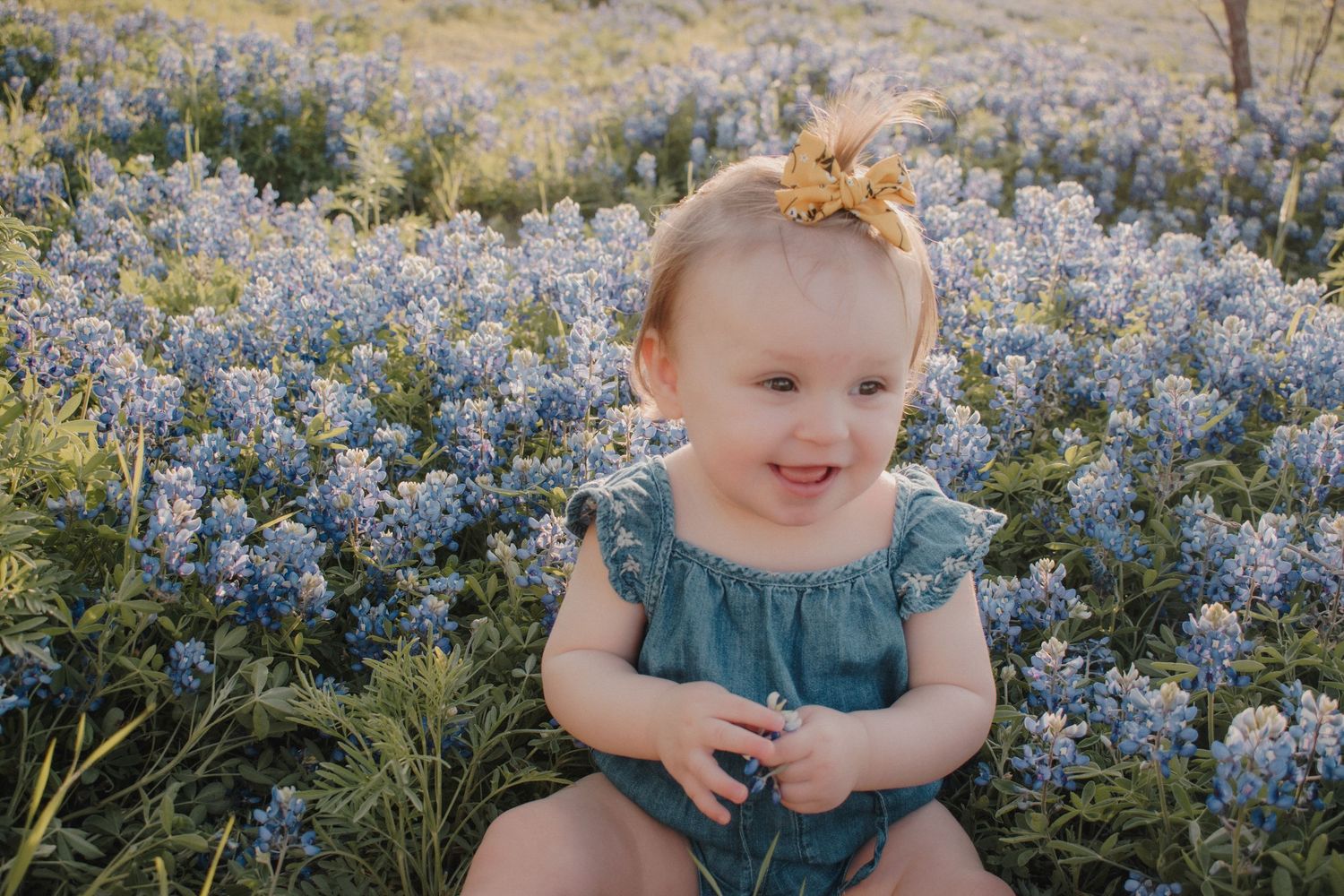 A happy baby girl in a teal romper sits among blooming blue wildflowers outdoors.