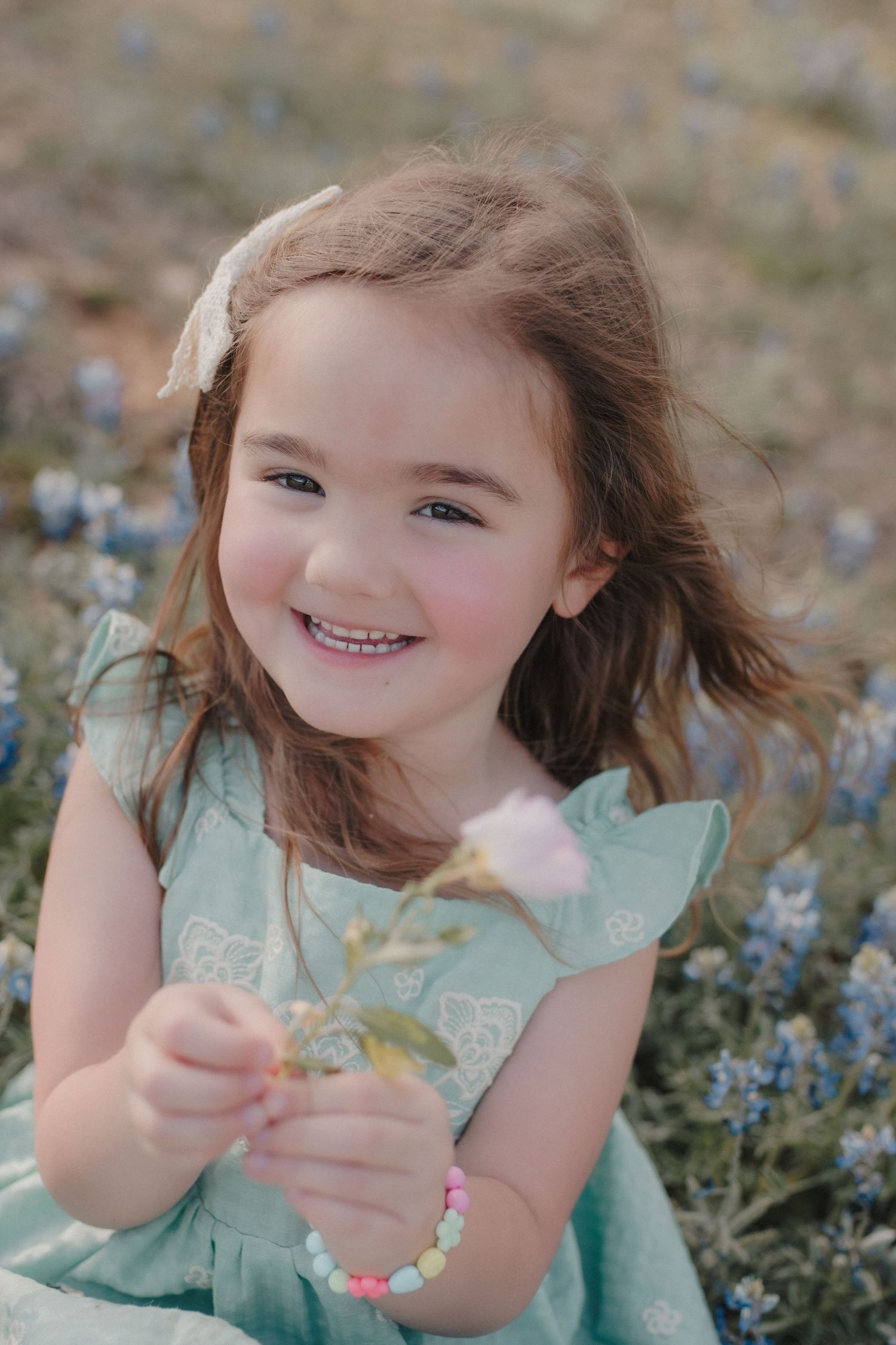 Smiling little girl in mint dress holding a pink flower in a bluebonnet field with a bow in her hair.
