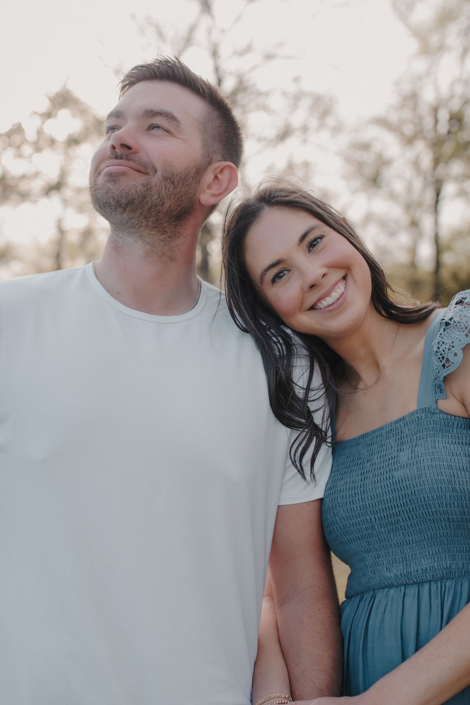 A smiling couple outdoors at golden hour, woman in teal dress leaning on man in white tee, trees in background.