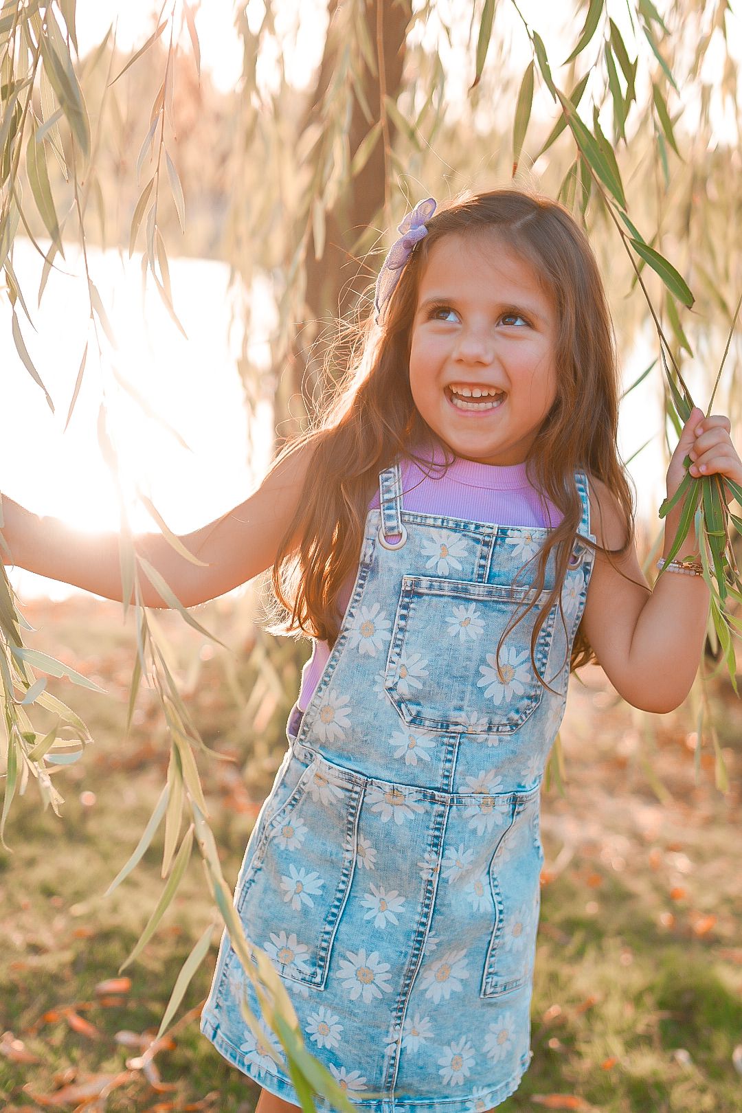 Happy young girl in denim floral jumper plays among weeping willow branches near lake during sunset portrait session.