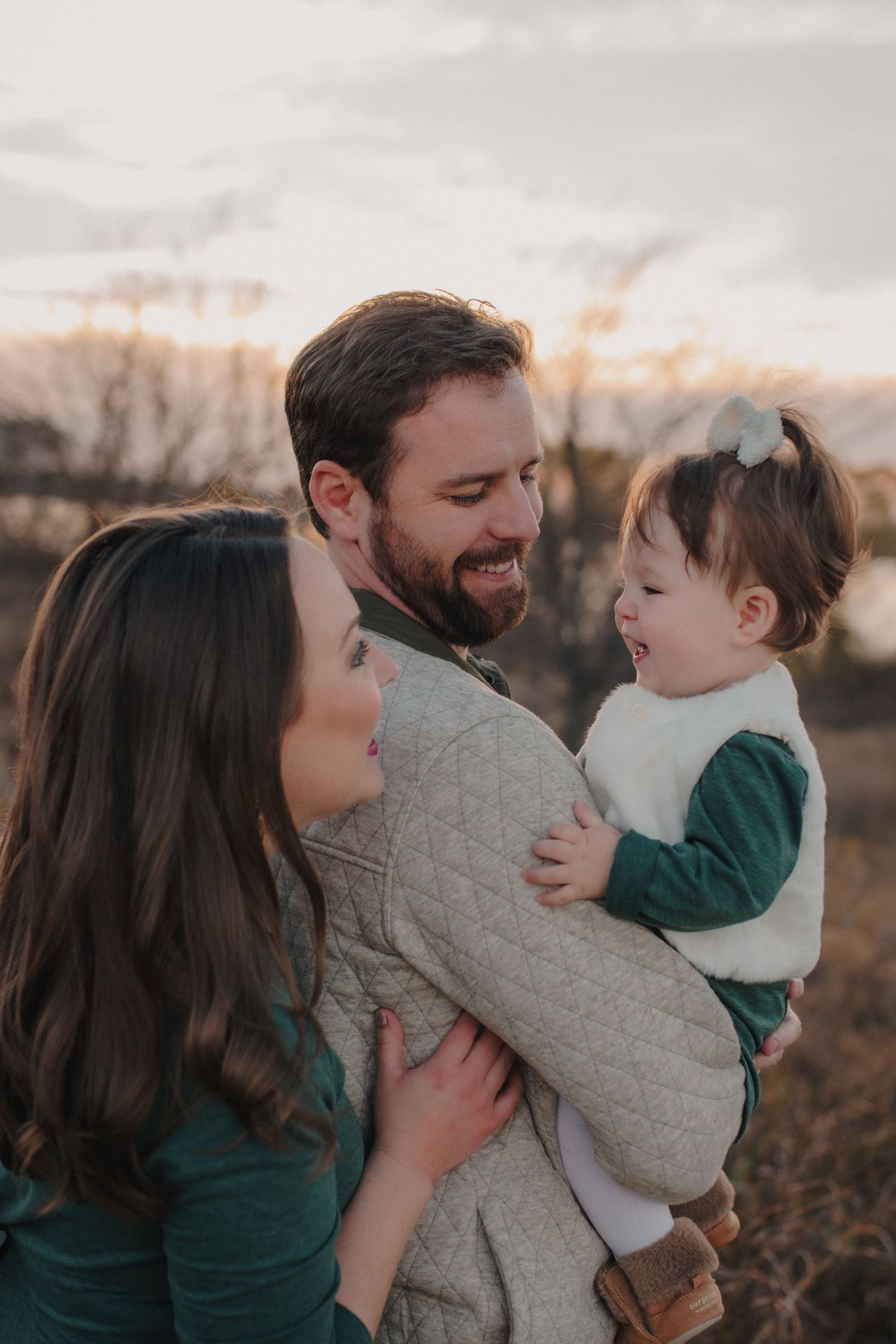 Happy young family with bearded father holding laughing toddler girl outdoors at golden sunset.