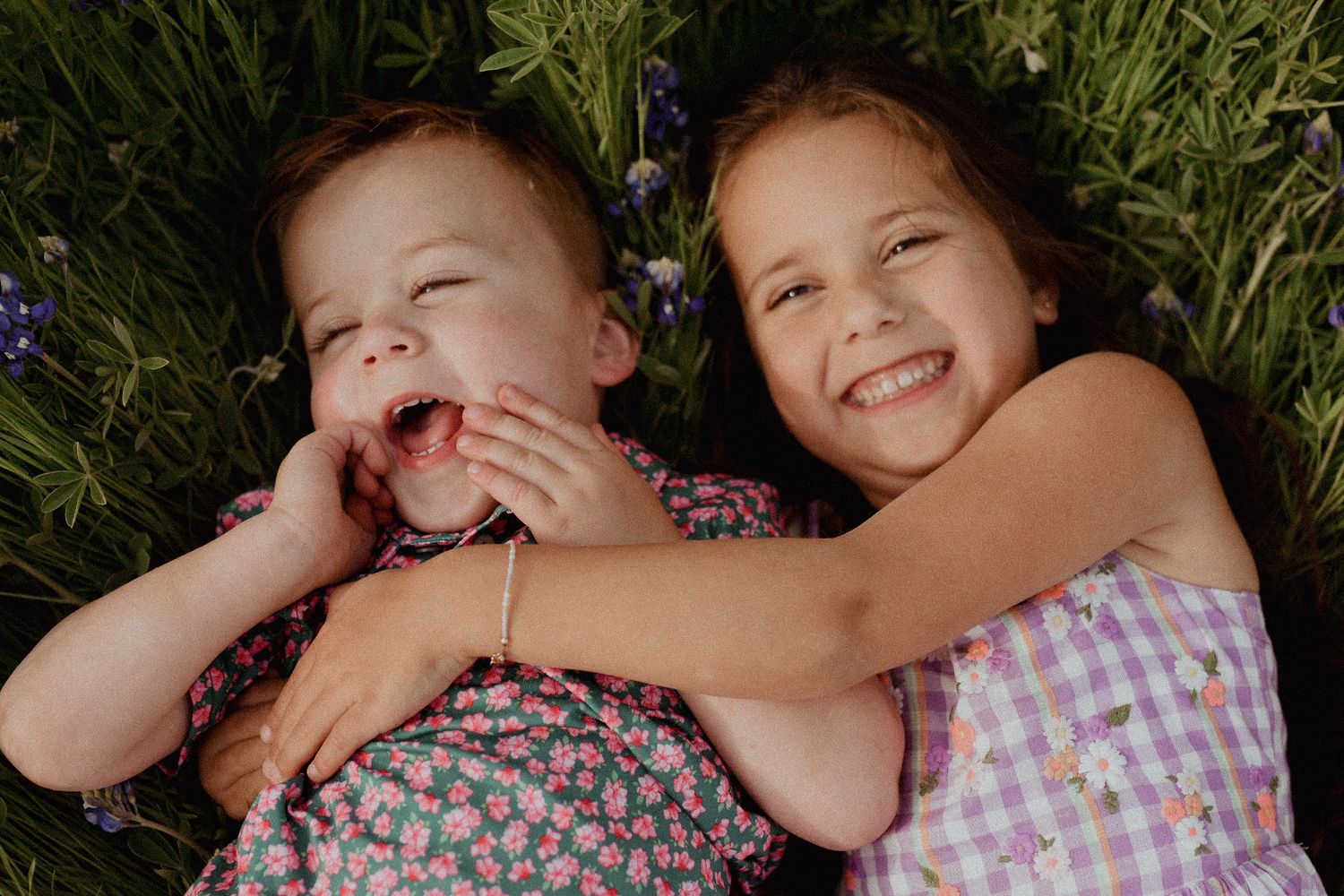 Two young siblings laugh and smile together while lying in a field of green grass and flowers.