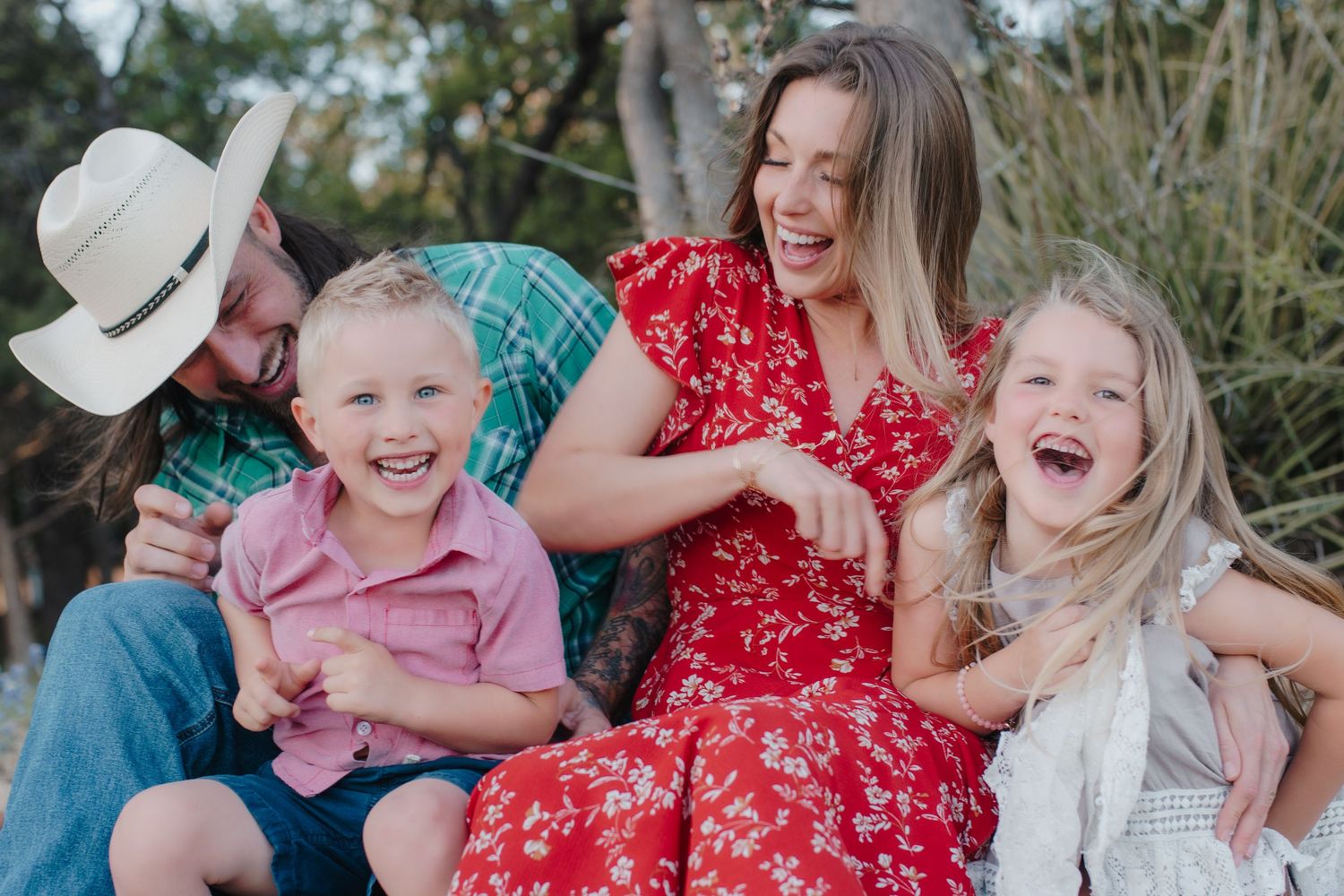 Happy family of four laughing together outdoors, parents with young boy and girl in red floral dress and cowboy hat.