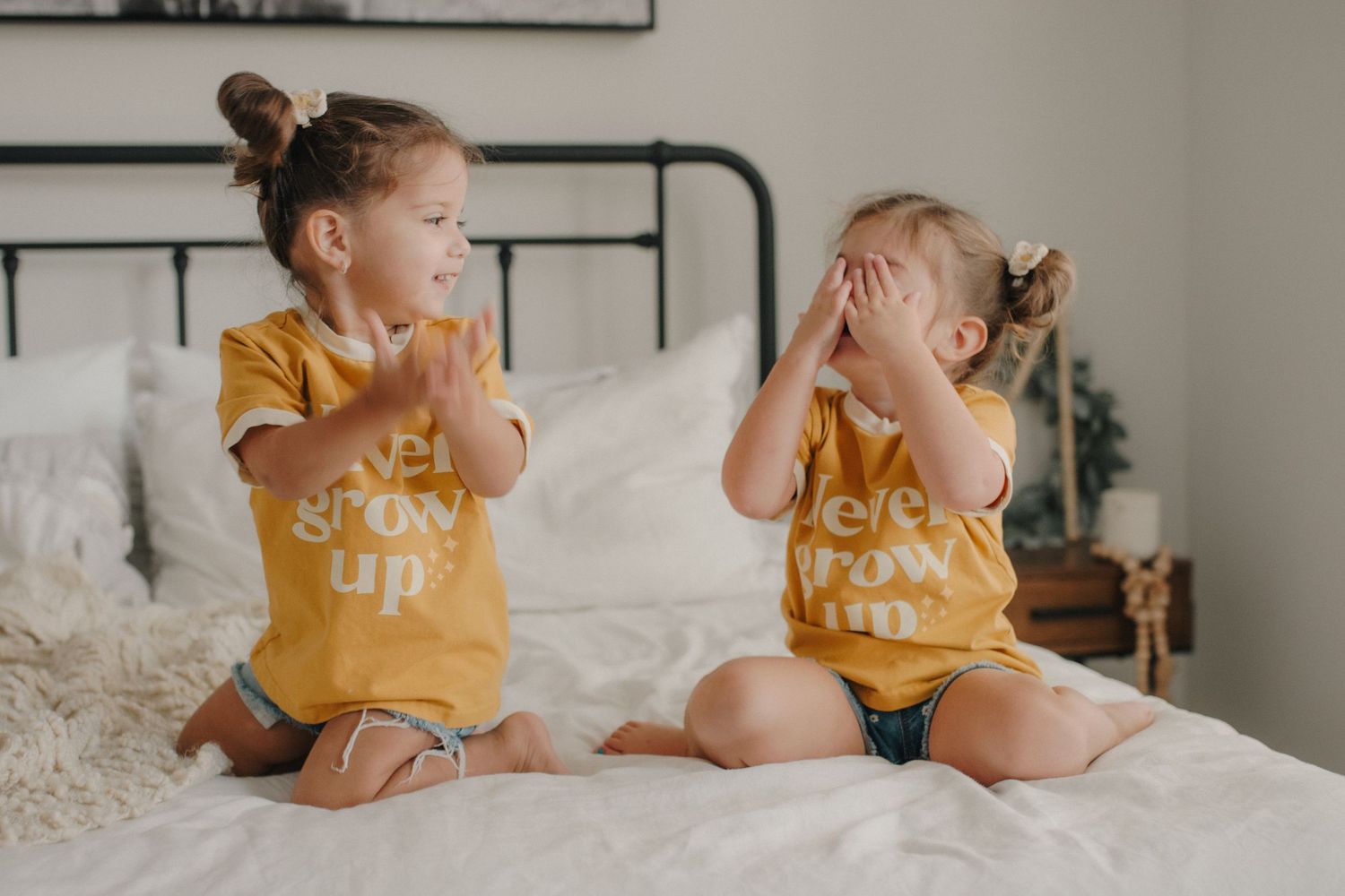 Two young sisters in matching yellow t-shirts sitting on a white bed, one clapping and one covering her face.