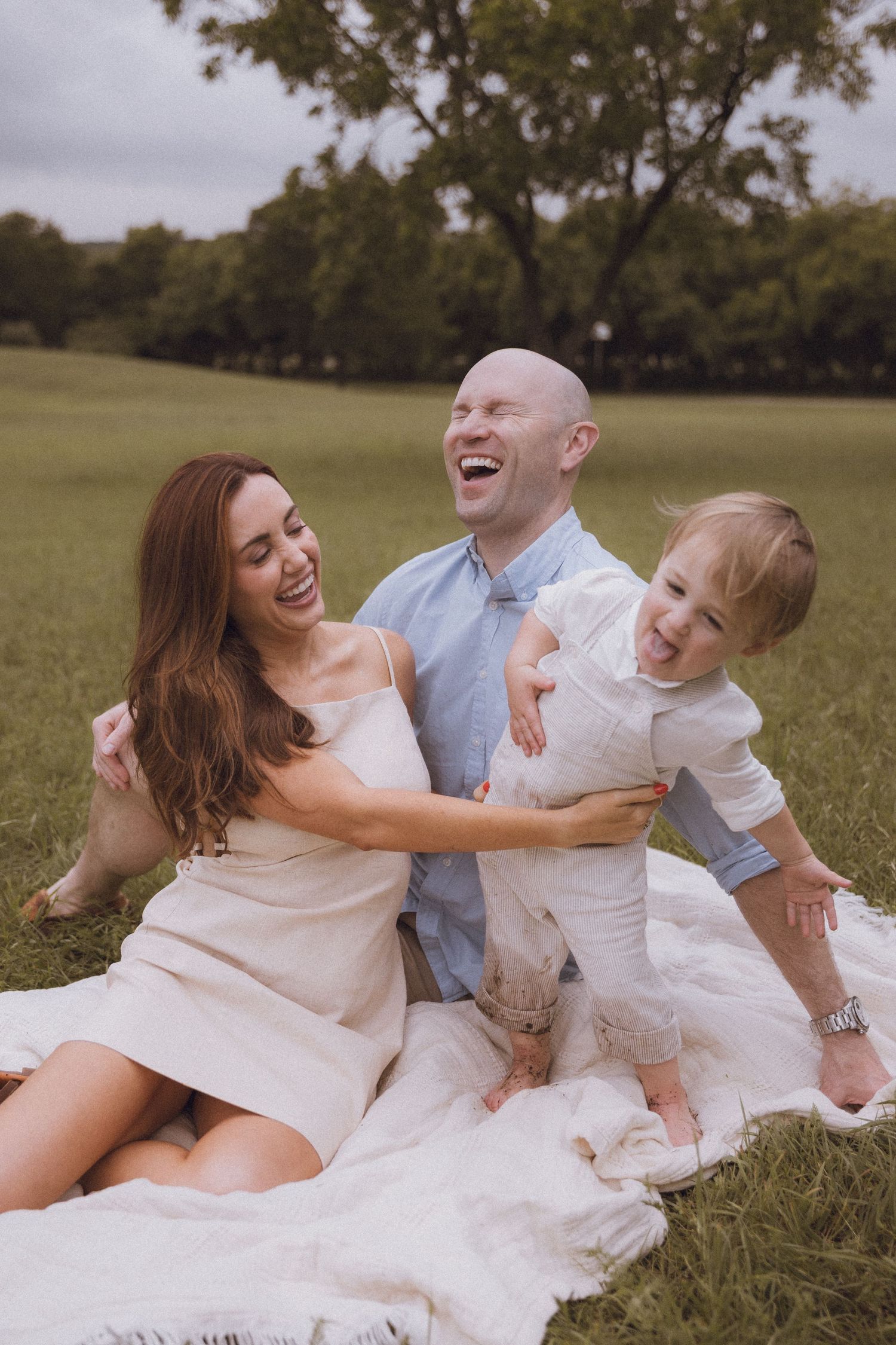 Happy family laughing together on a blanket in a green park, parents and toddler enjoying a sunny outdoor picnic.