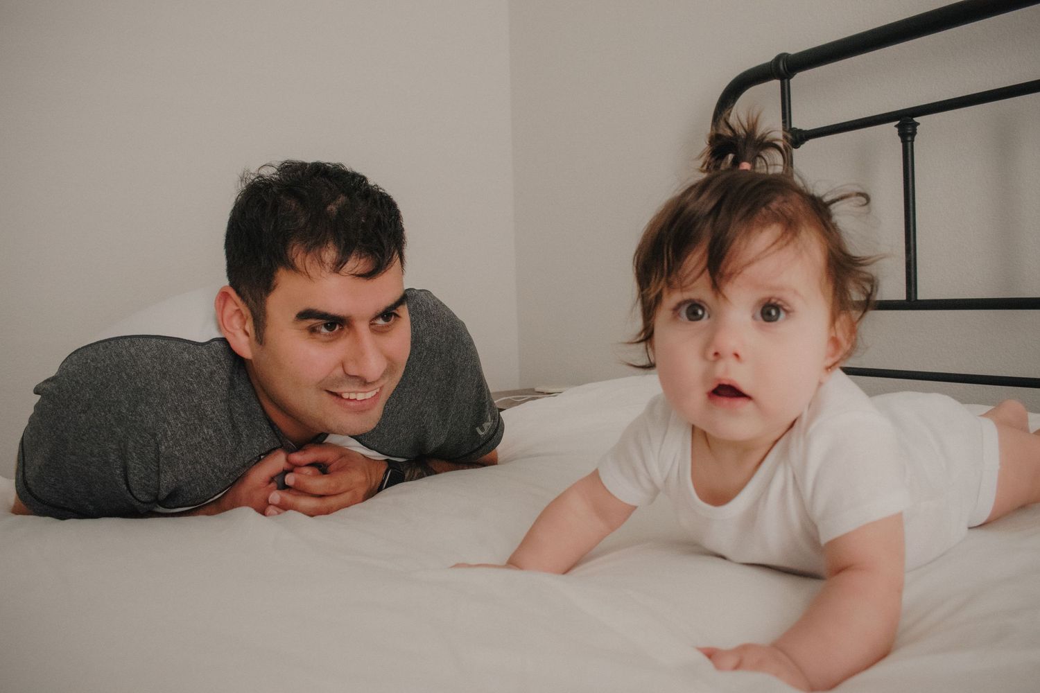 A father lies on a bed smiling beside his curious wide-eyed baby in a white onesie.
