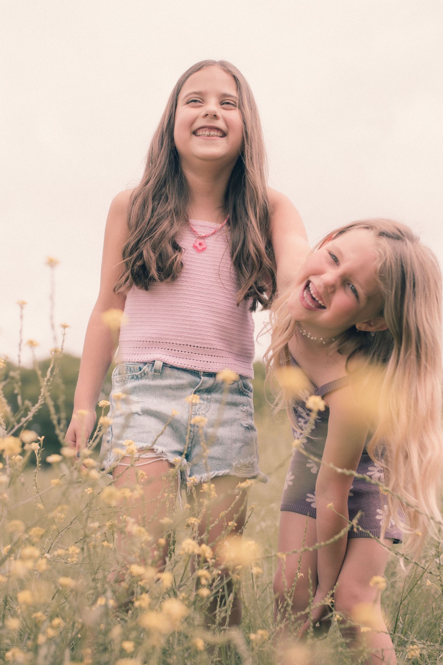 Two laughing girls enjoy a sunny day in a wildflower meadow, one in a pink top and denim shorts, the other in a floral top.