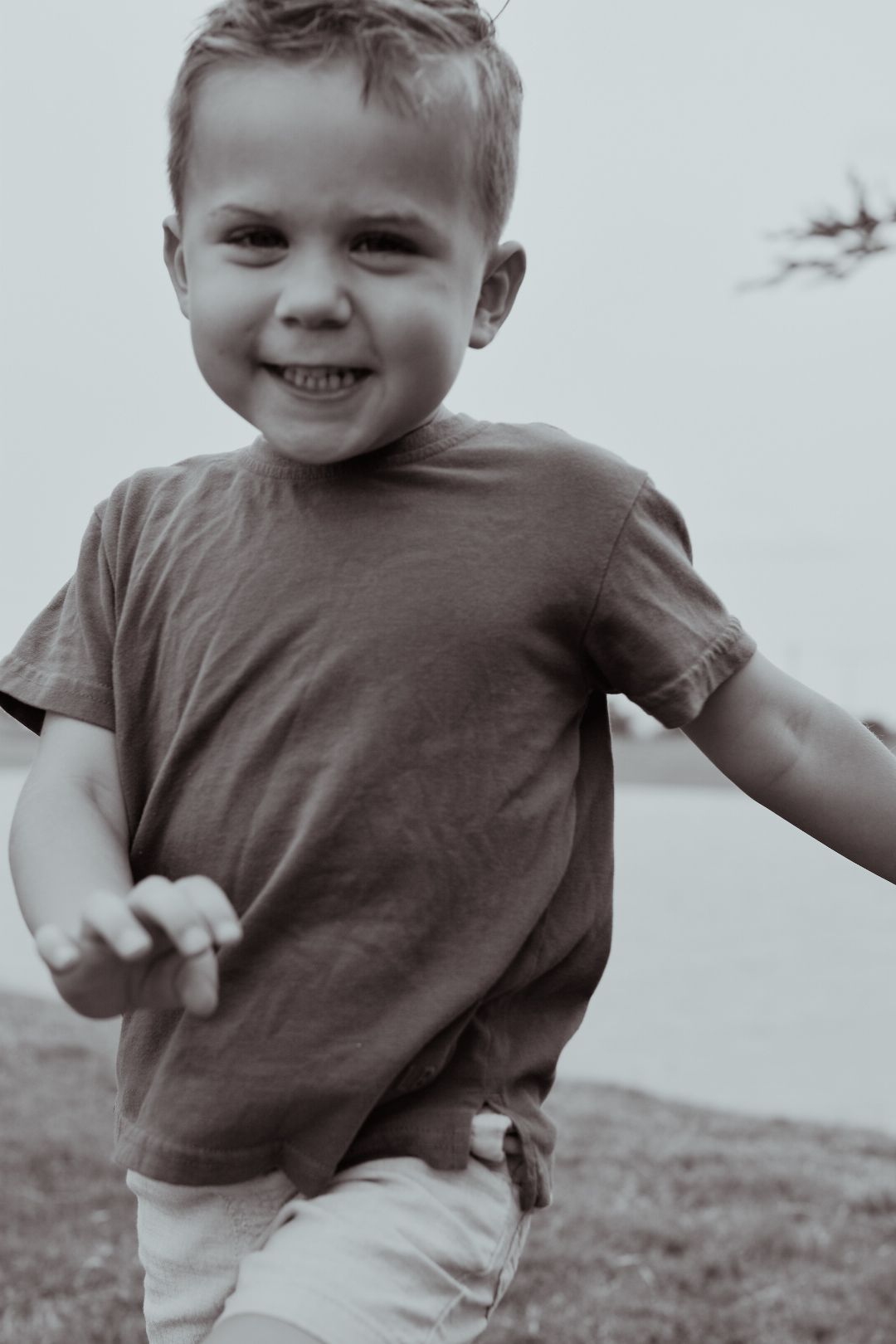 Smiling young boy in gray t-shirt runs outdoors near water in black and white toned children portrait photo.