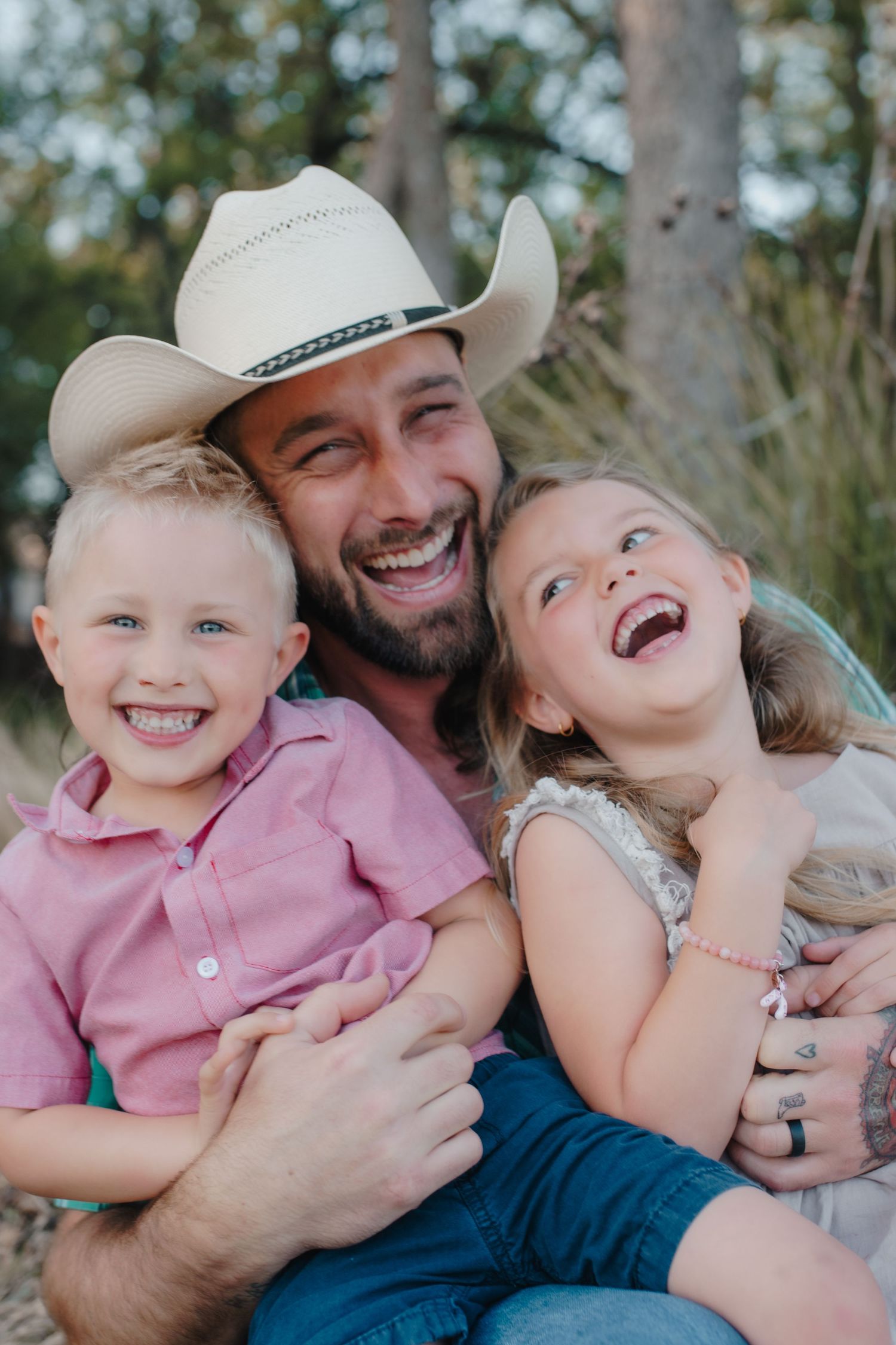A bearded man in a cowboy hat laughs joyfully with two young children outdoors among trees.