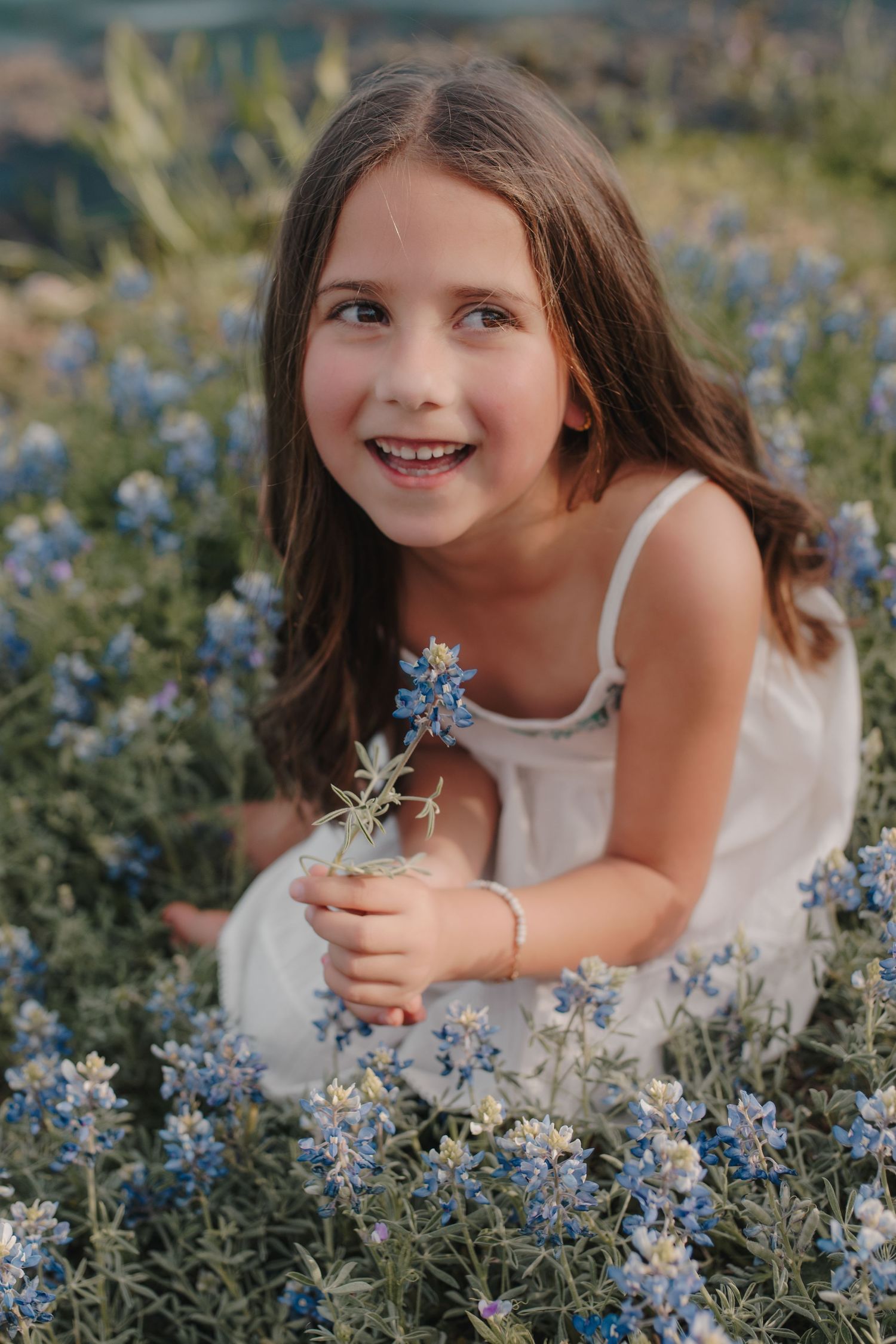 Happy young girl in a white dress sitting in a field of blue wildflowers, holding a small bouquet.
