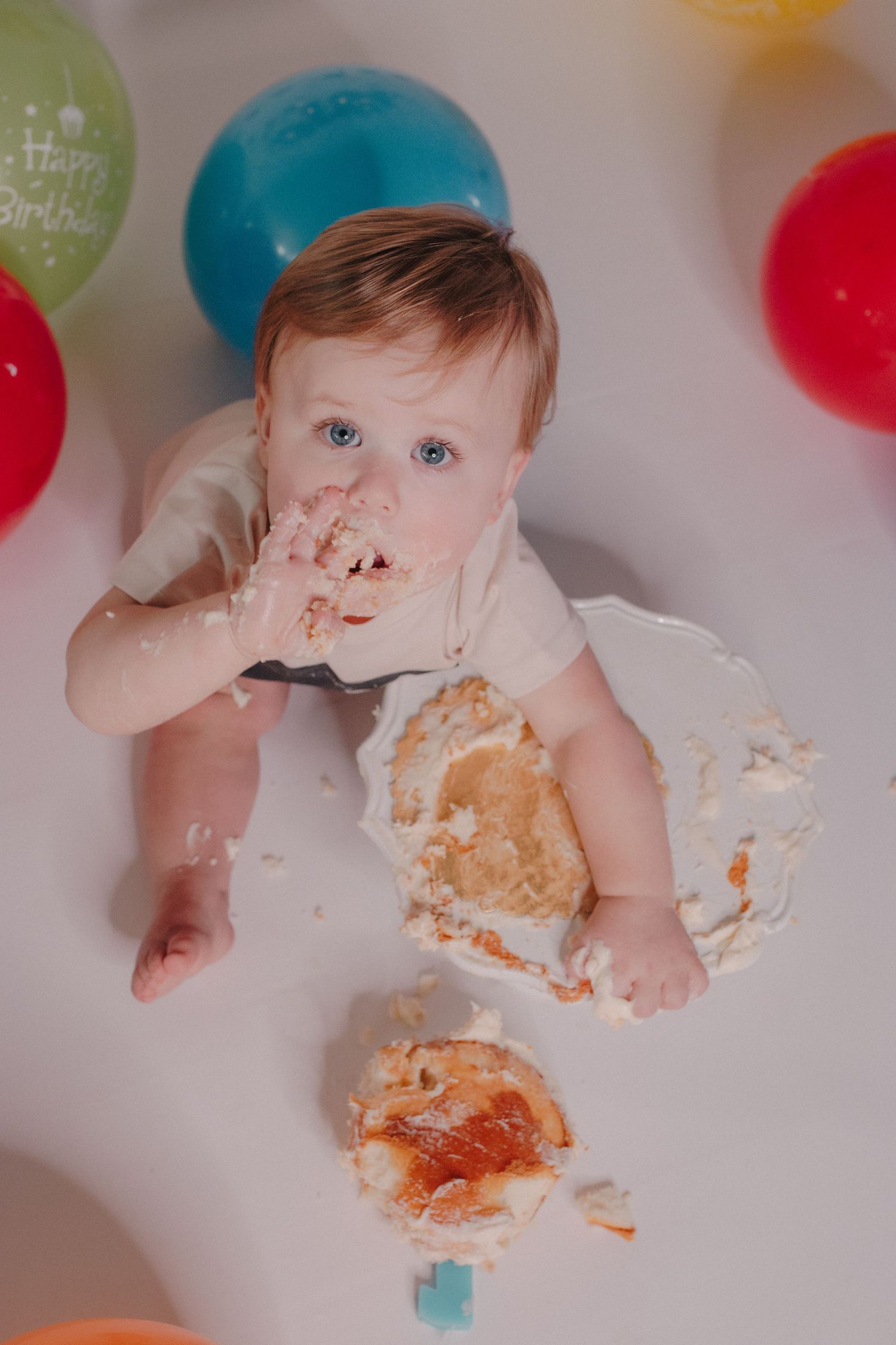 Happy blue-eyed baby celebrating first birthday smashing cake surrounded by colorful balloons.
