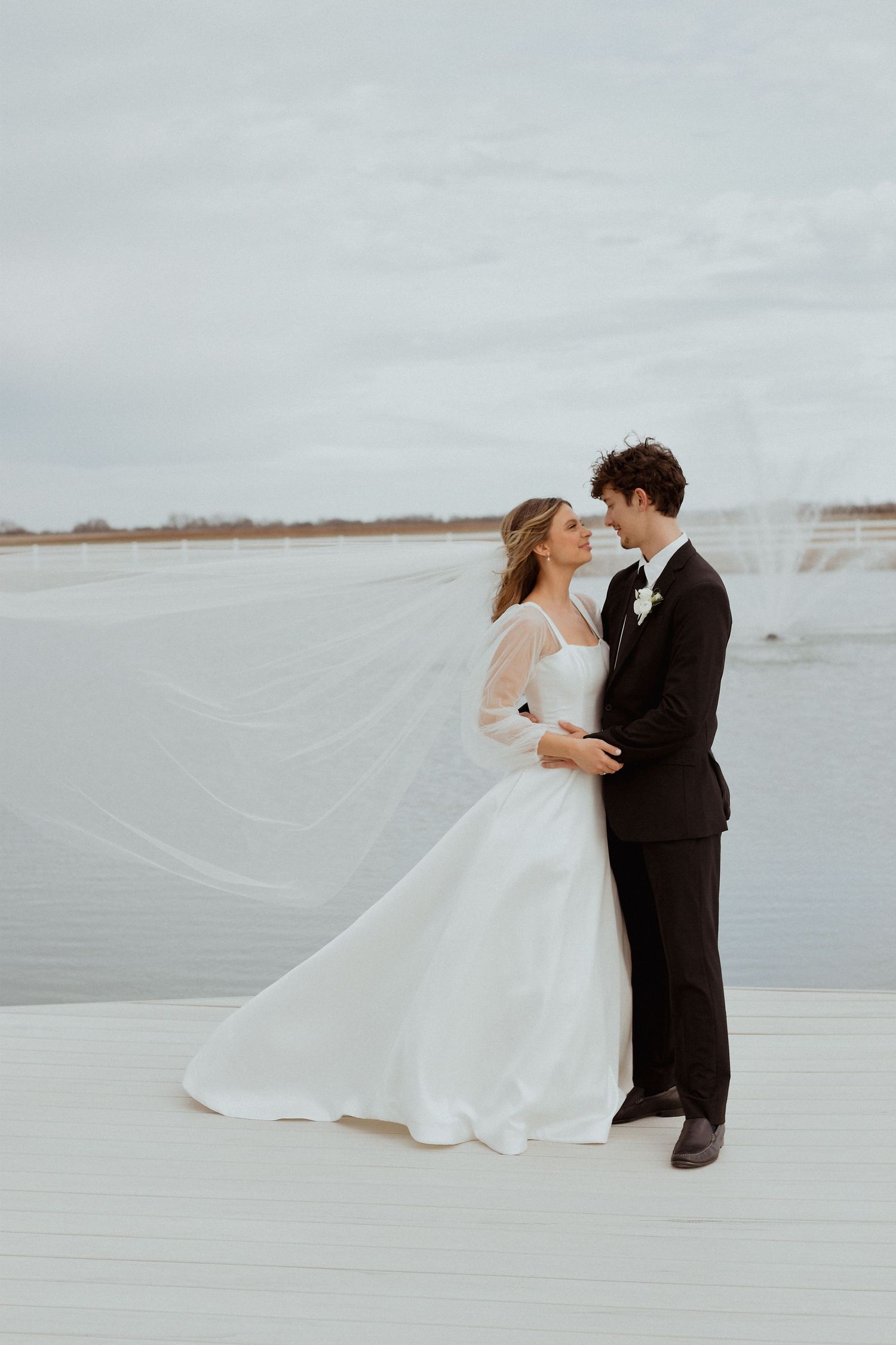 Bride in white gown with flowing veil embraces groom in black suit on dock with lake backdrop on cloudy day.