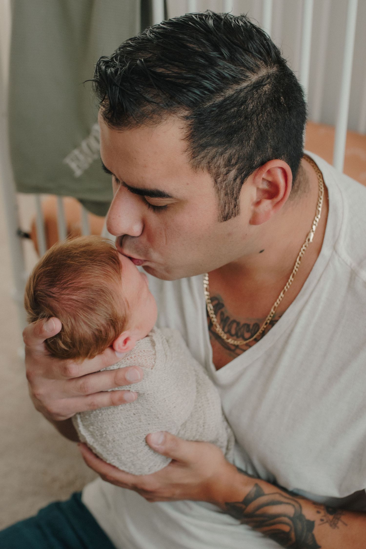 Tattooed father tenderly kissing his swaddled newborn baby while seated indoors.