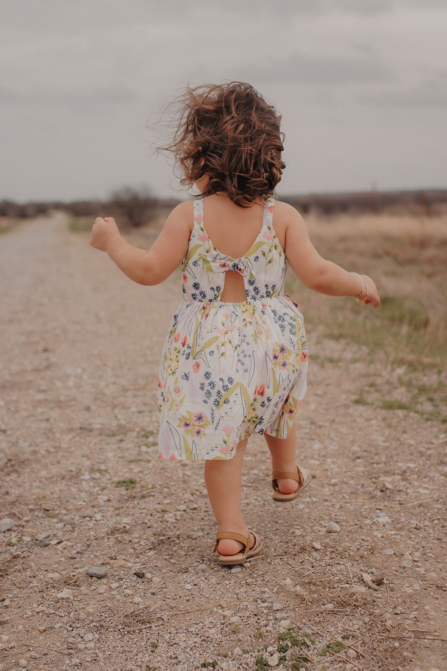 Curly-haired toddler girl in floral dress running down a dirt path outdoors.