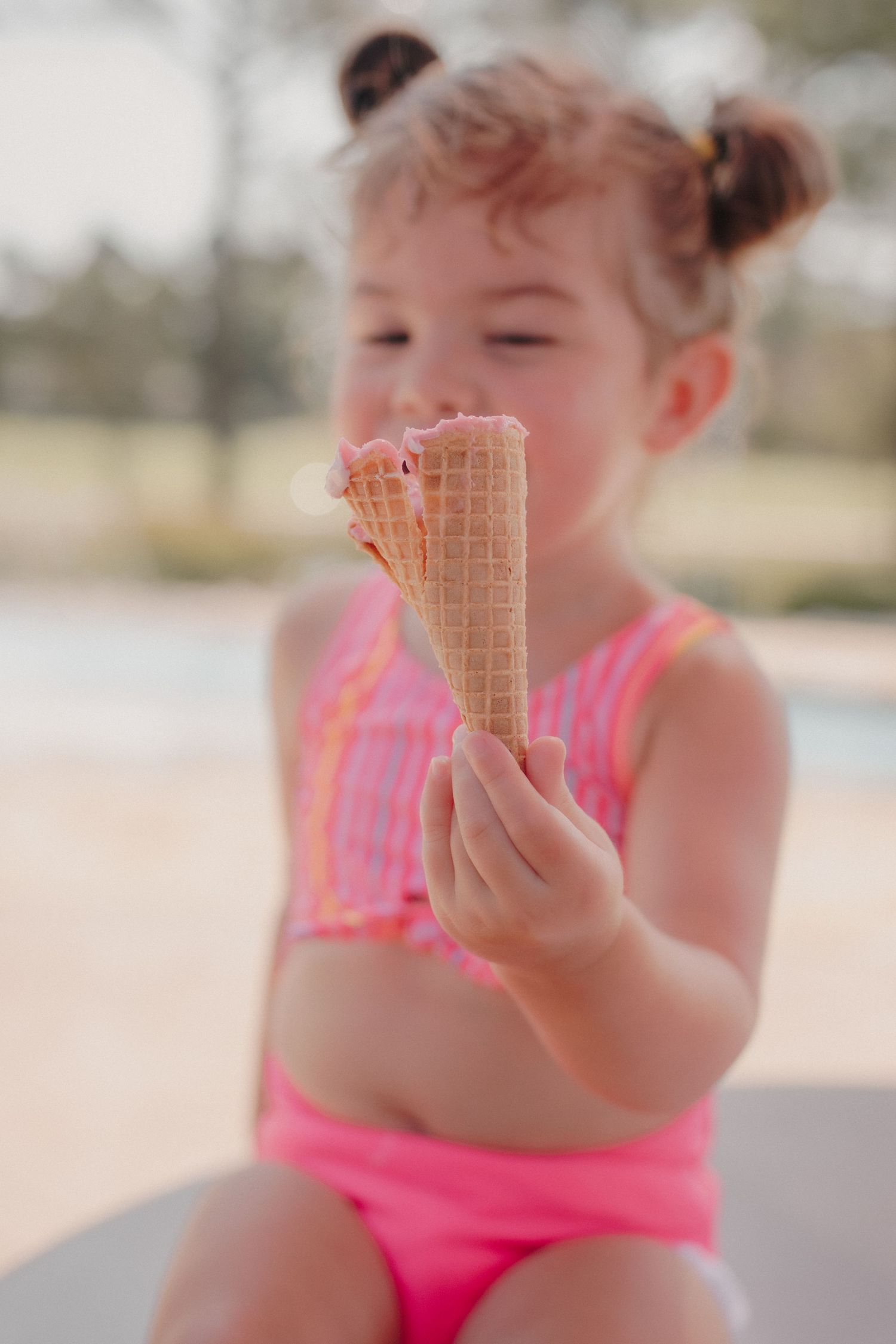 Cute toddler girl in pink swimsuit holding melting ice cream cone outdoors.