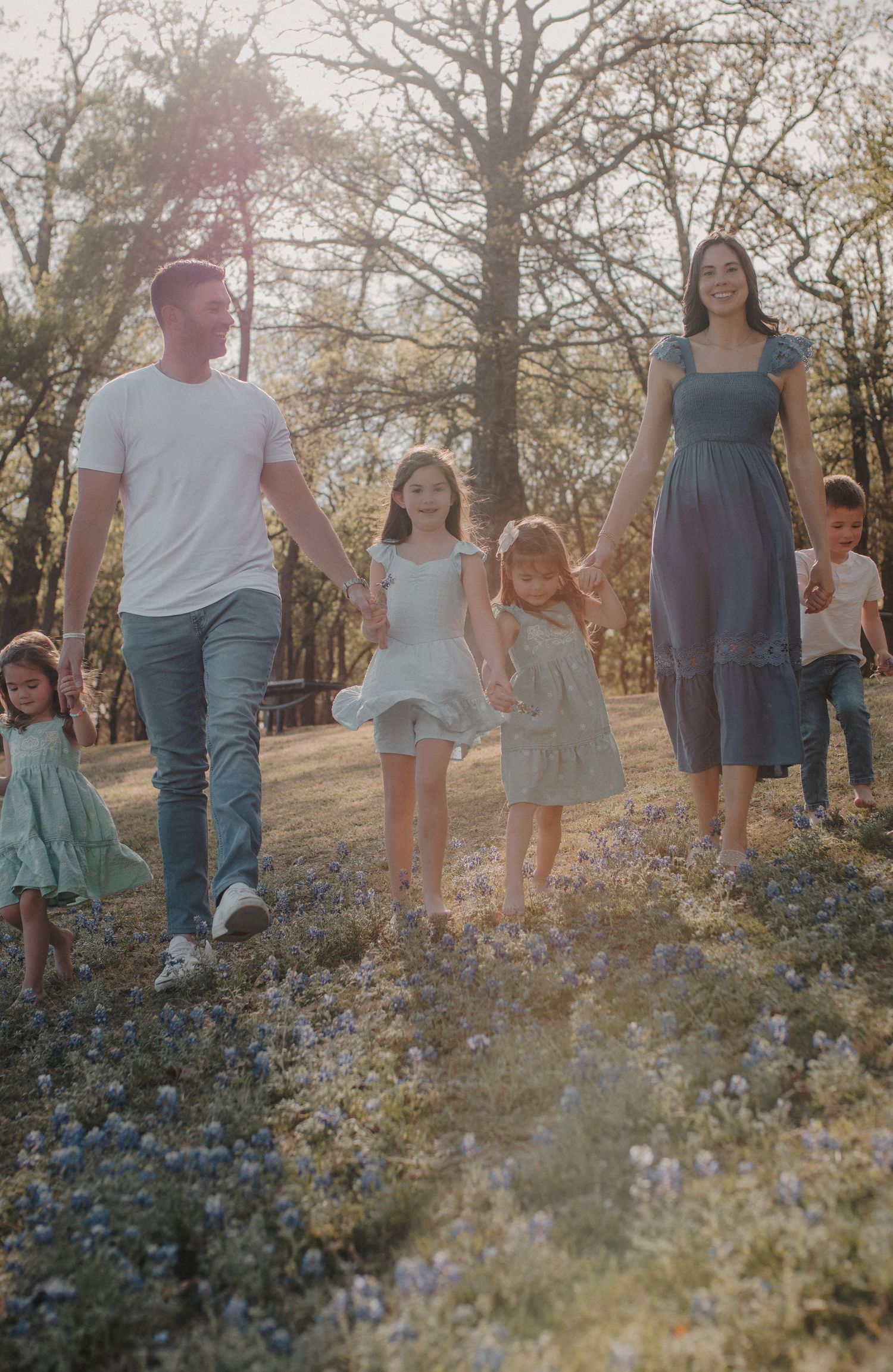 Family of five holding hands walking through a sunlit wildflower meadow surrounded by trees.
