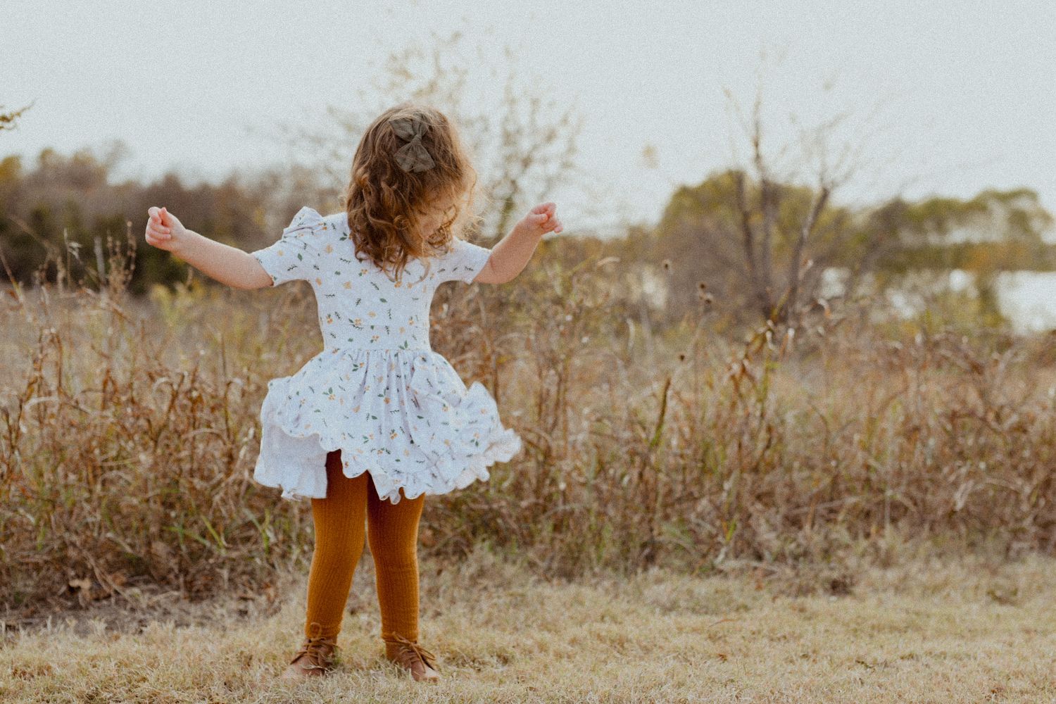 A young girl in a white floral dress and brown boots twirls in a golden autumn field.