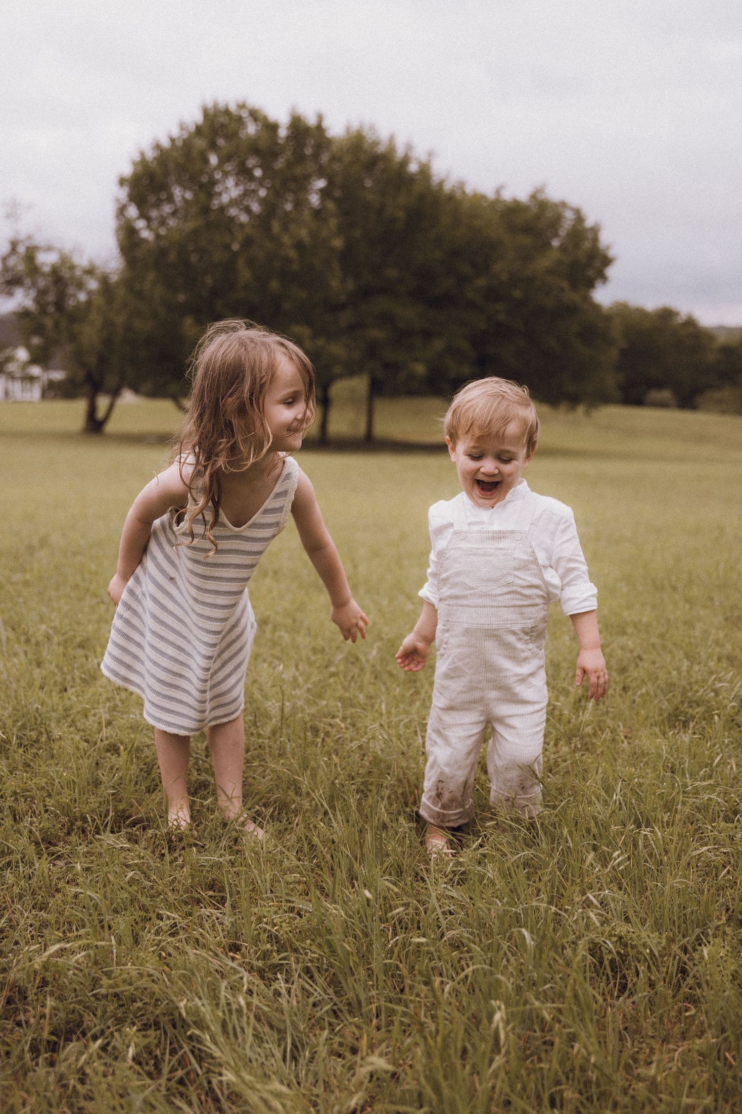 Two happy young children playing in a green grassy field, girl in striped dress and toddler boy in white overalls.