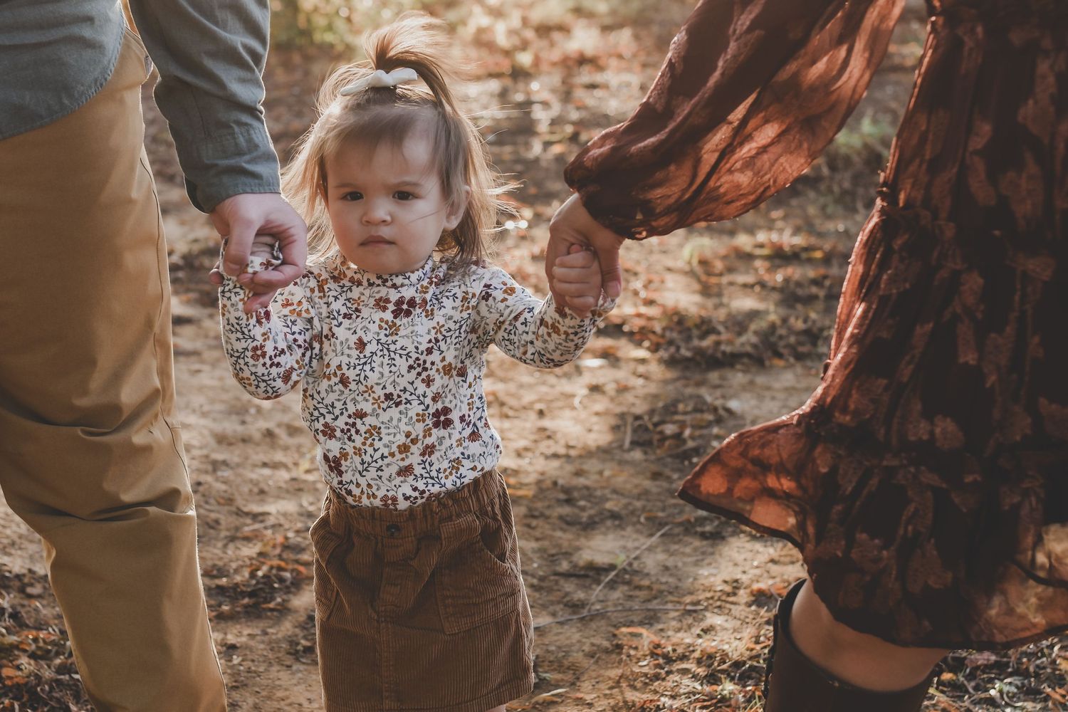Toddler girl in floral shirt and brown skirt holds hands with parents during outdoor fall family portrait session.