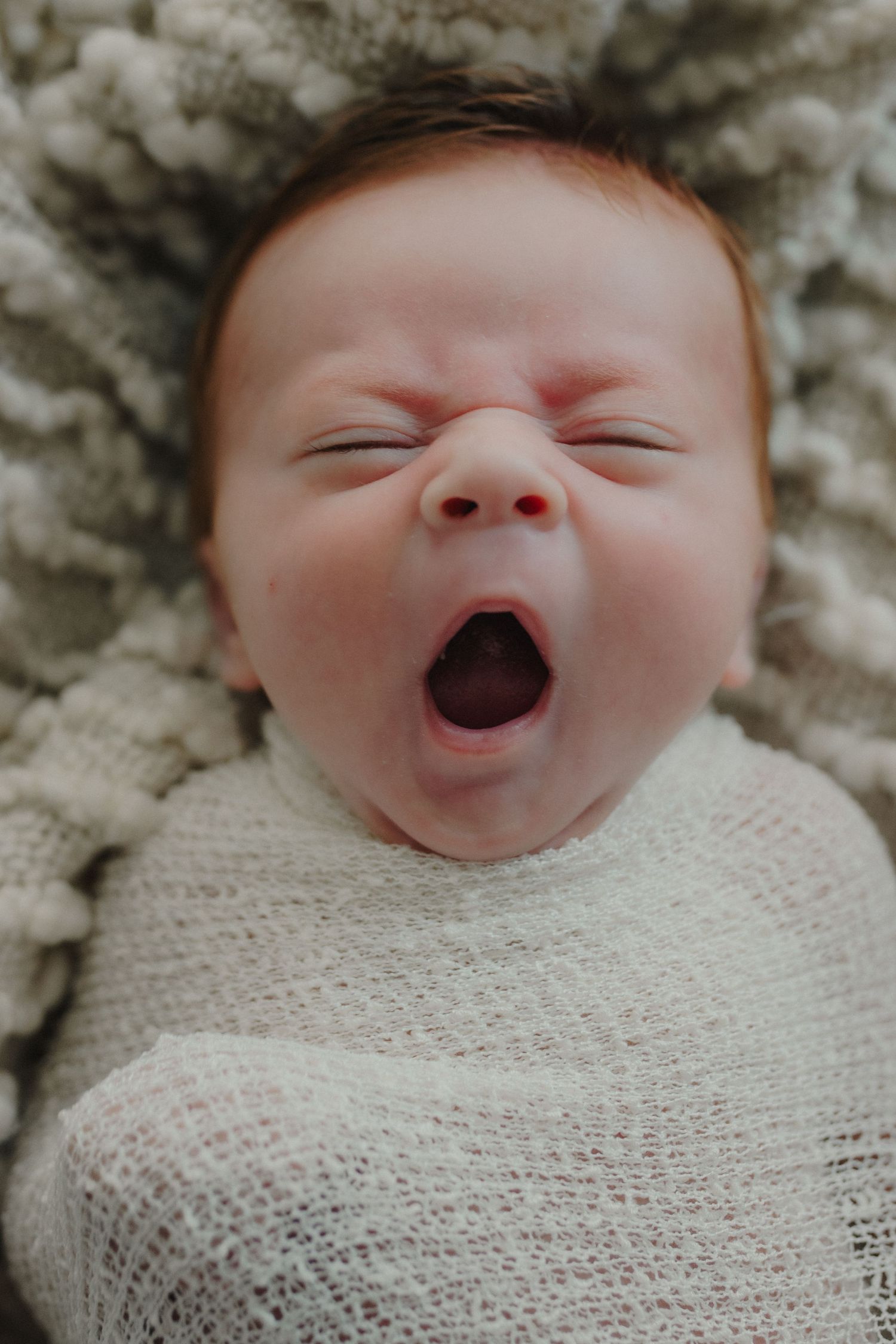 Peaceful newborn baby yawning while swaddled in white wrap on a soft knit blanket.