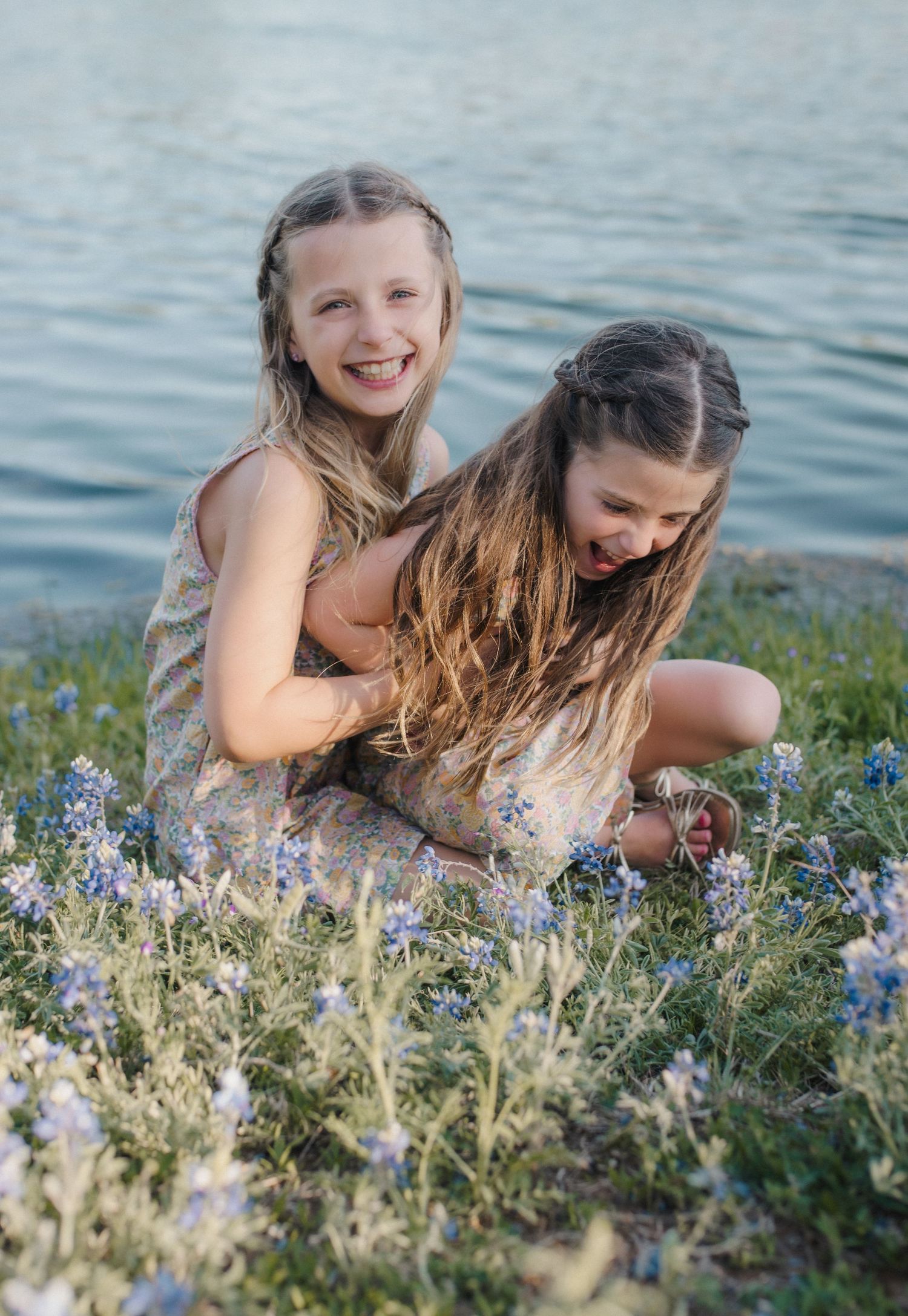 Two young girls laughing and hugging among bluebonnets by a lake, wearing floral dresses with braided hair.