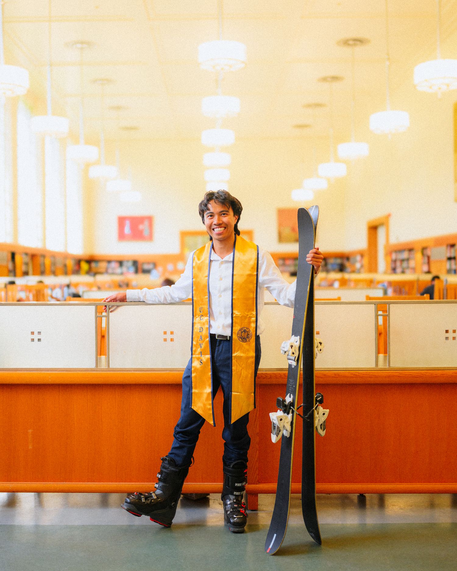 Graduate wearing gold stole poses with skis in university library setting.