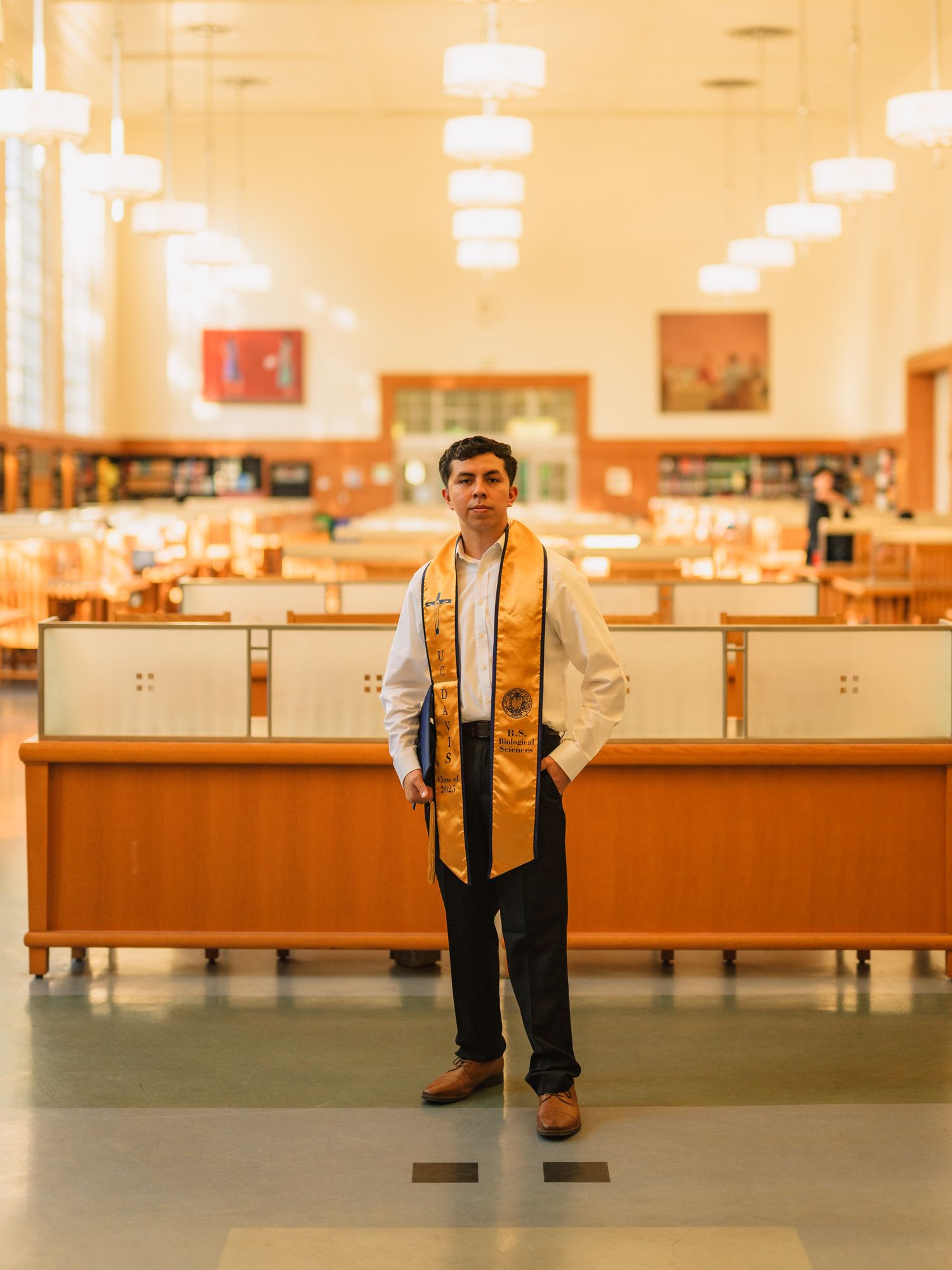 Graduate in formal attire and yellow stole poses in an indoor library setting.