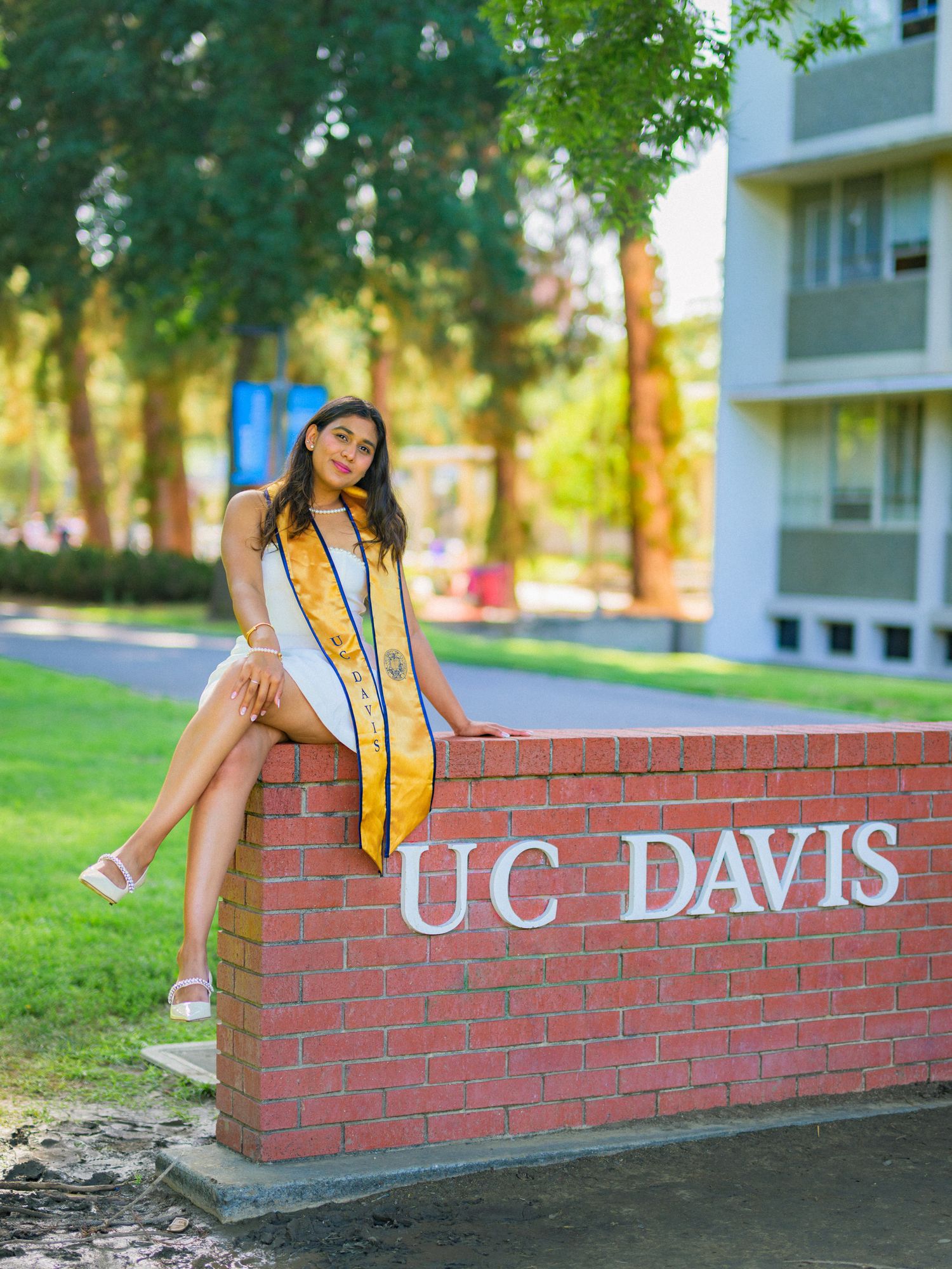 Graduate in white dress sits on UC Davis brick sign wearing graduation stole.