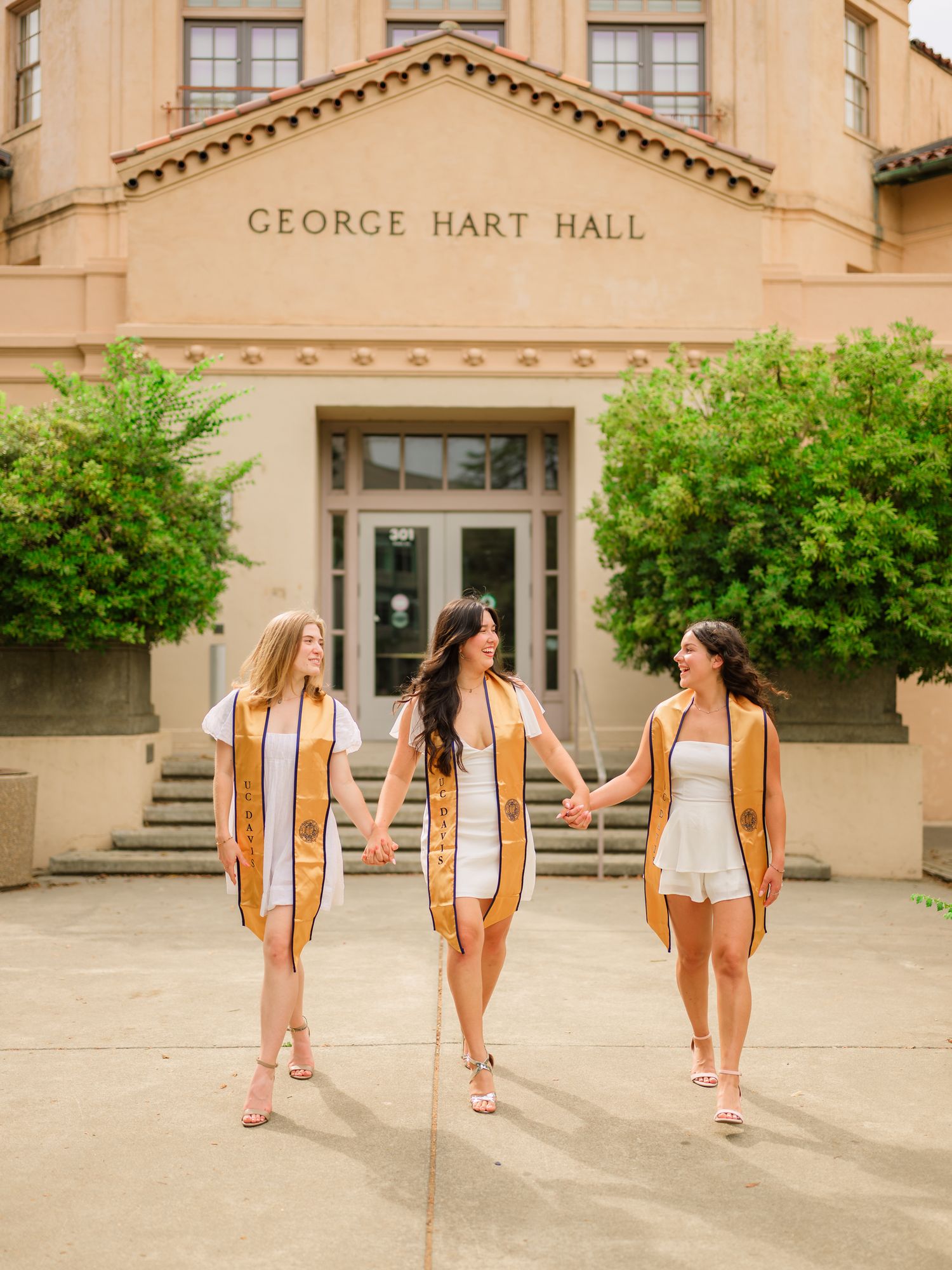 Three graduates in white dresses and gold stoles walk away from George Hart Hall during graduation photos.