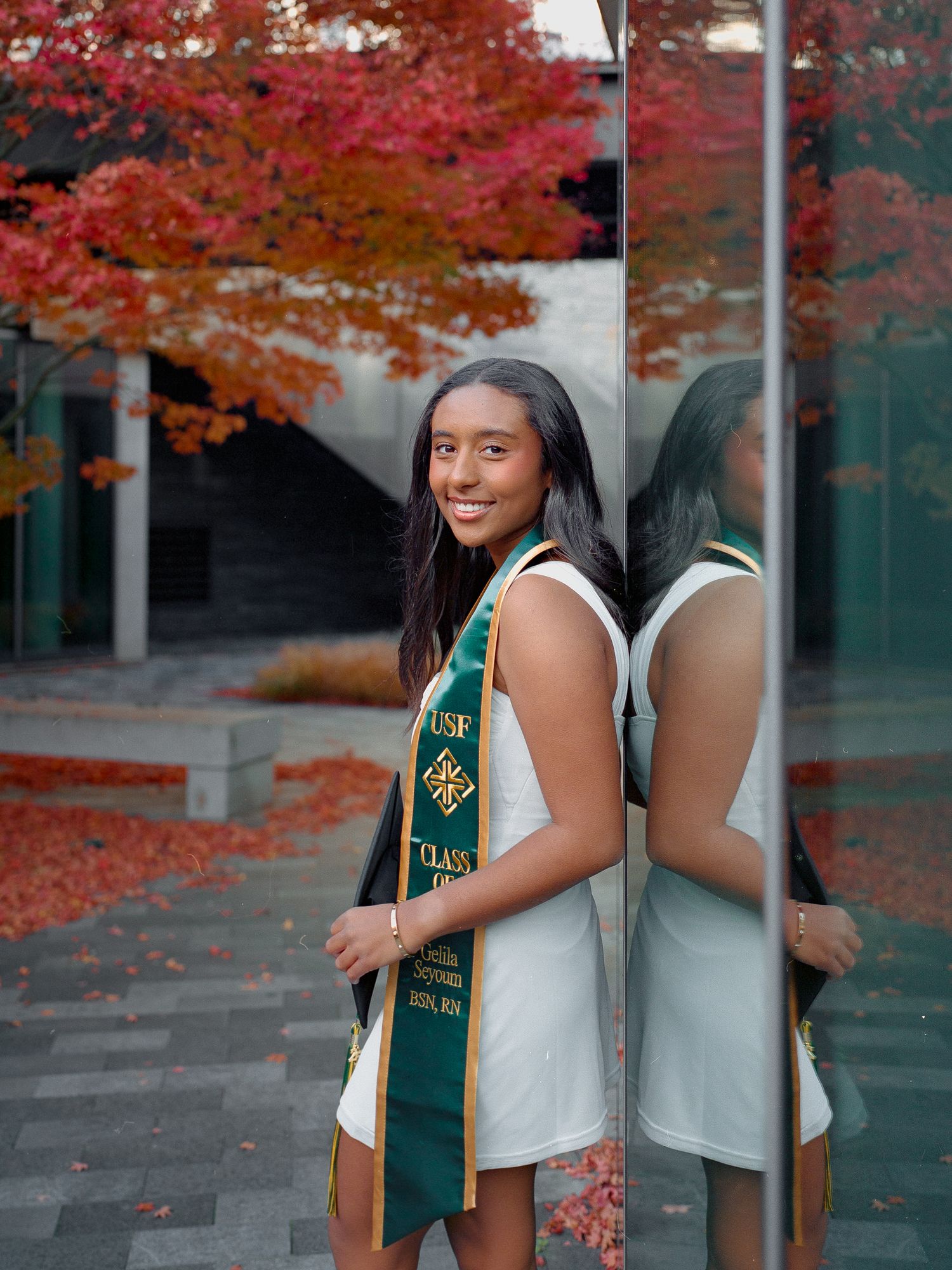 Graduate in green stole poses against autumn foliage and glass building backdrop.