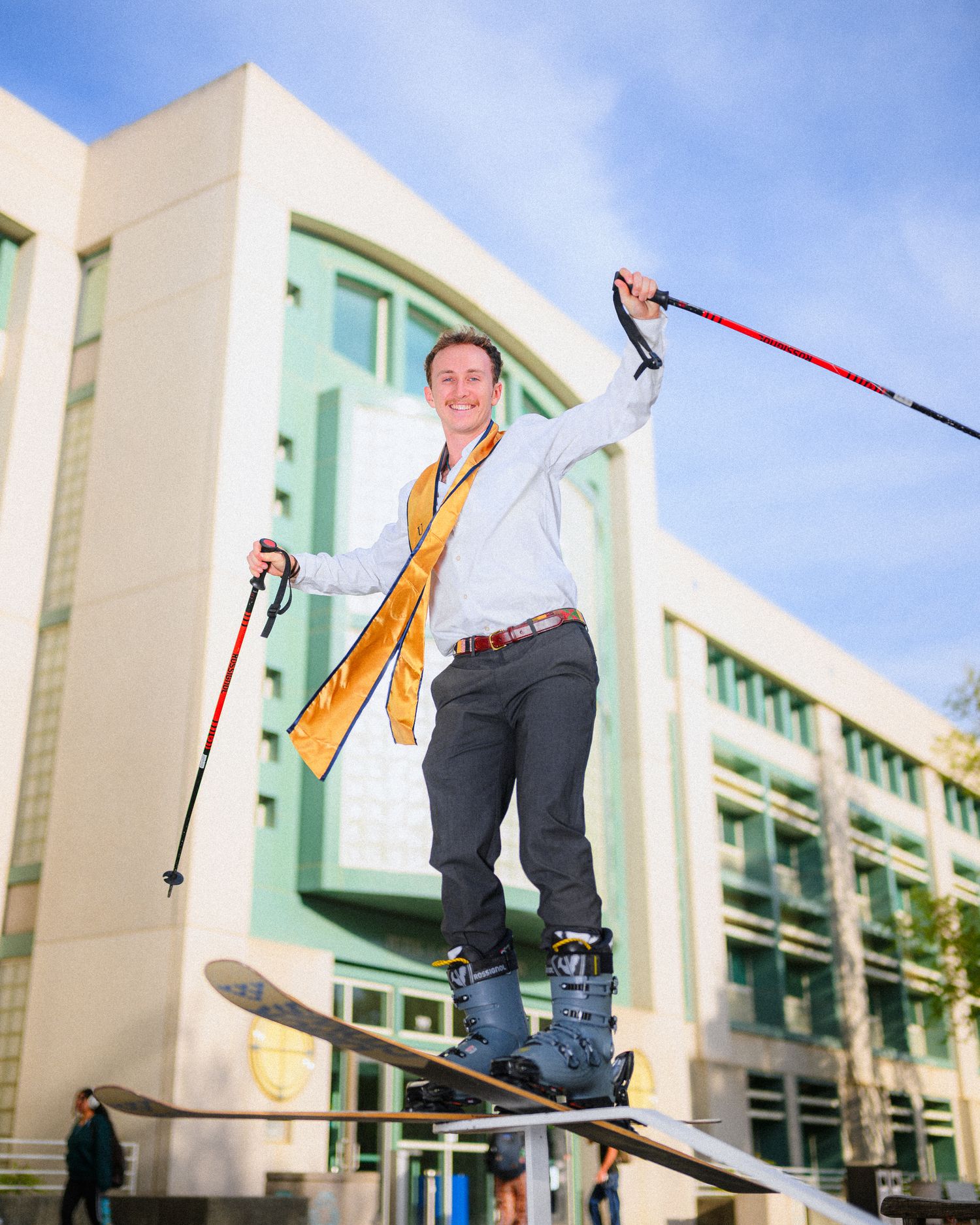 Graduate in ski gear poses with poles while balancing on skis outside campus building.