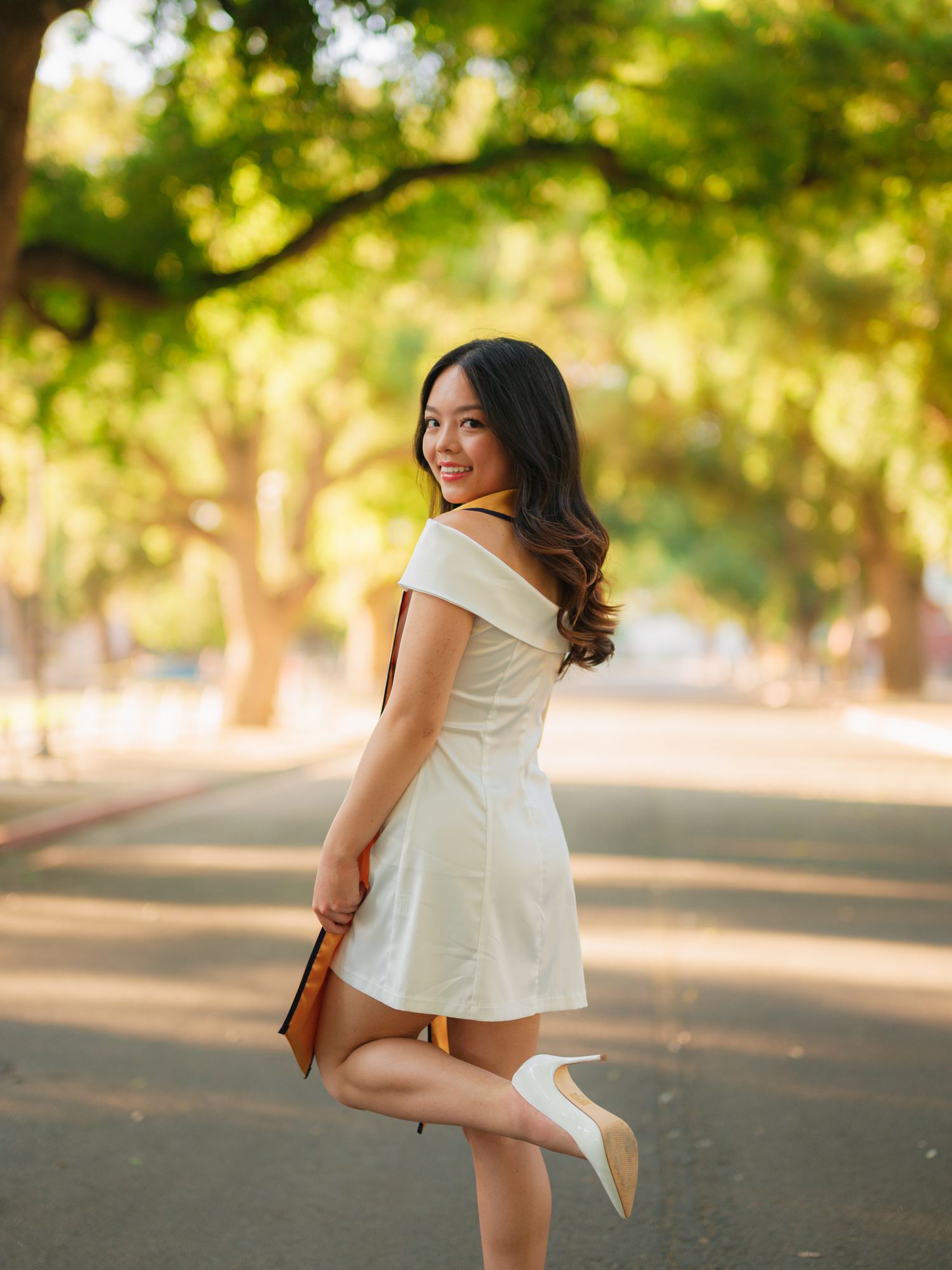 A person in a white dress and heels poses playfully on a tree-lined path with warm sunlit bokeh in the background.