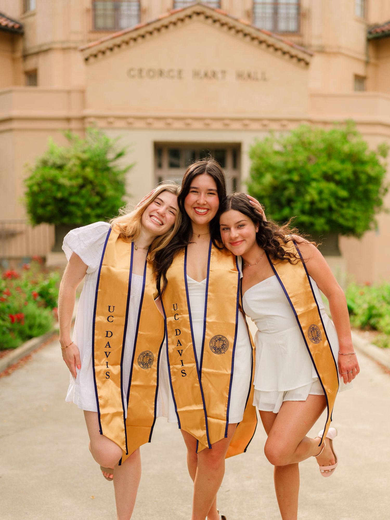 Three graduates in gold stoles pose playfully in front of a beige campus building.