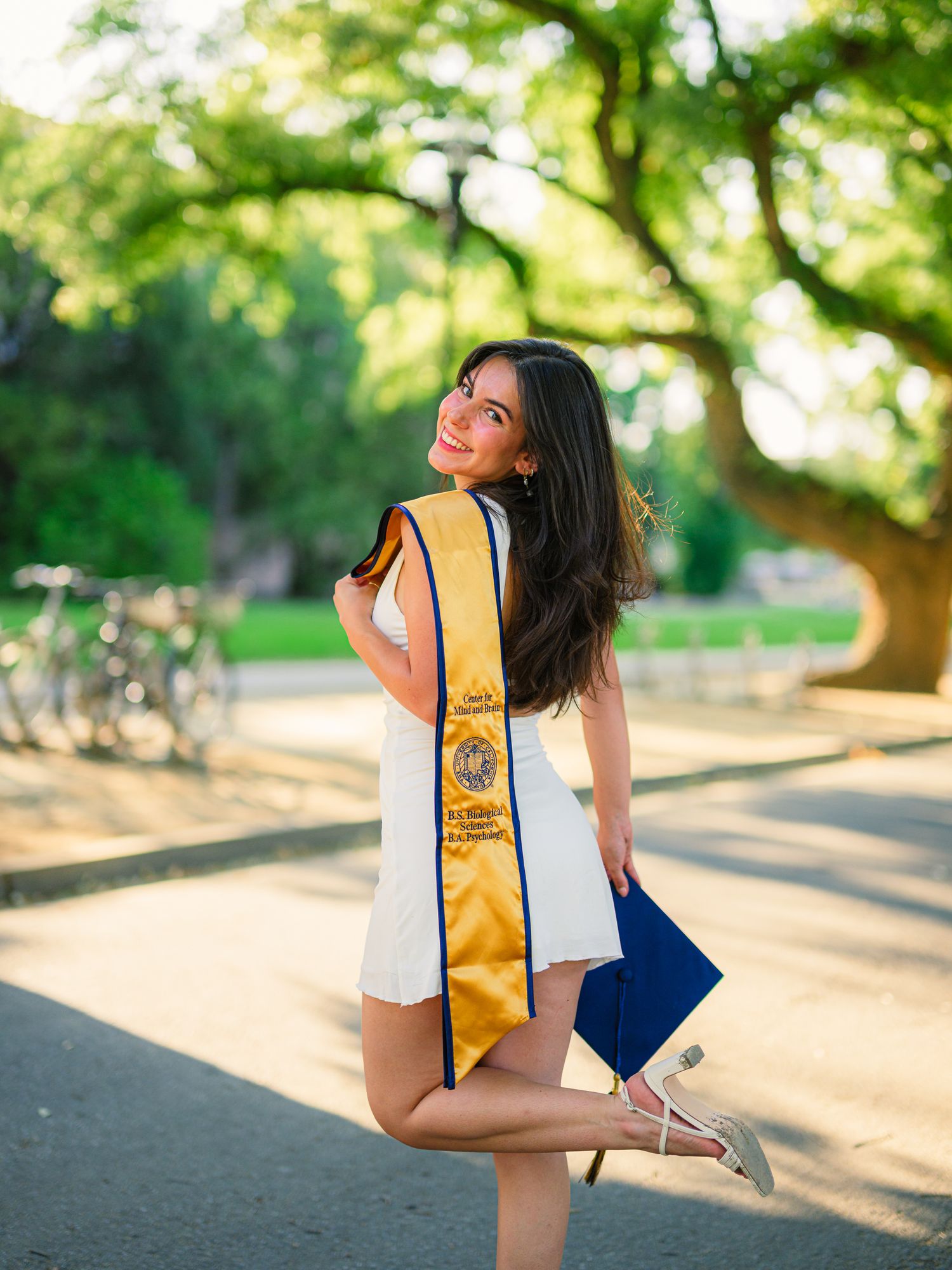 Graduate in white dress poses playfully with diploma on campus pathway.