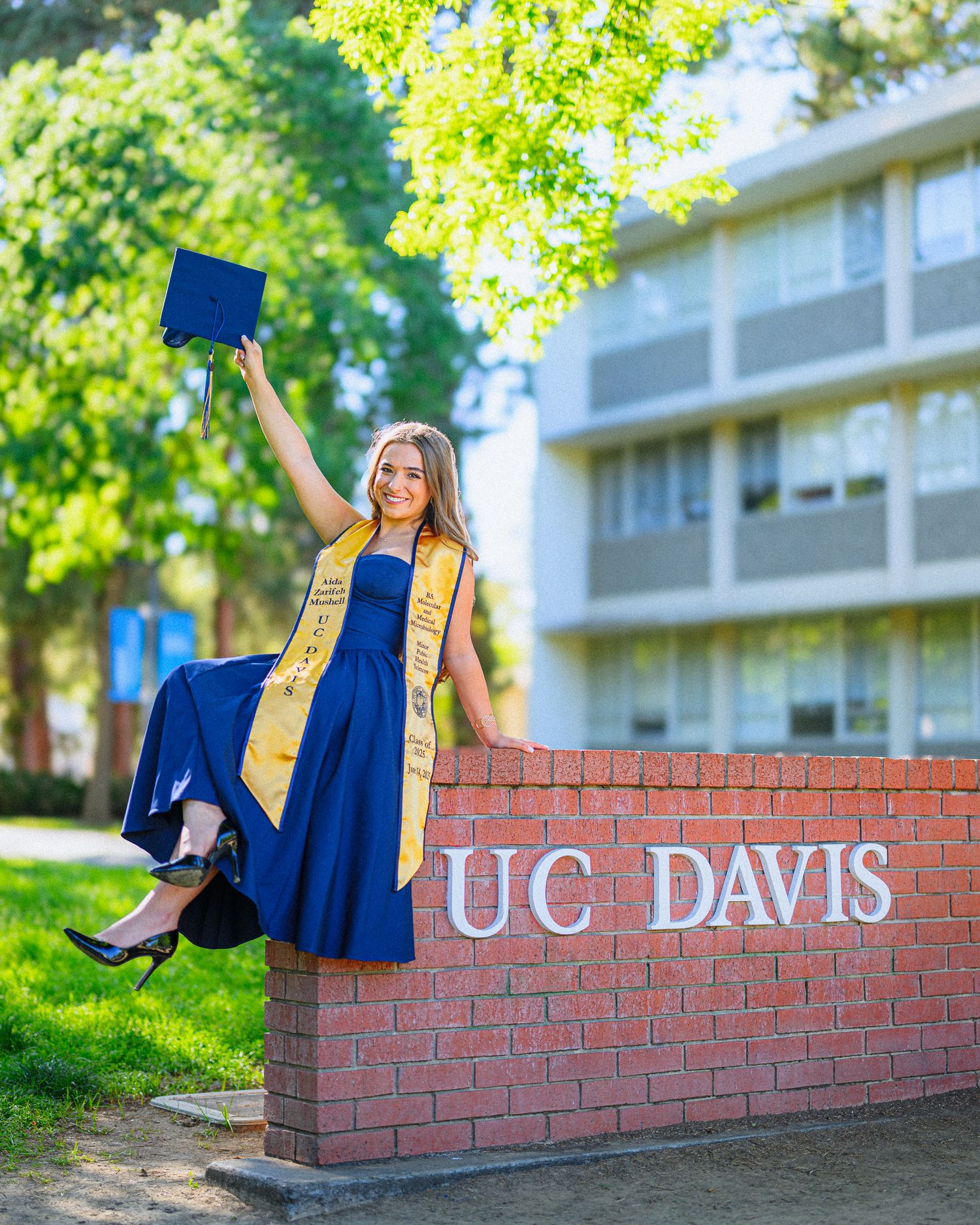 Graduate in blue regalia poses playfully on UC Davis brick wall during sunny graduation photoshoot.