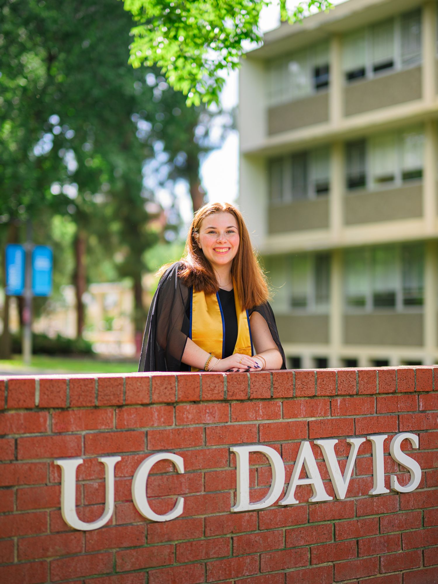 Graduate posing next to UC Davis brick sign in academic regalia with yellow stole on sunny day.