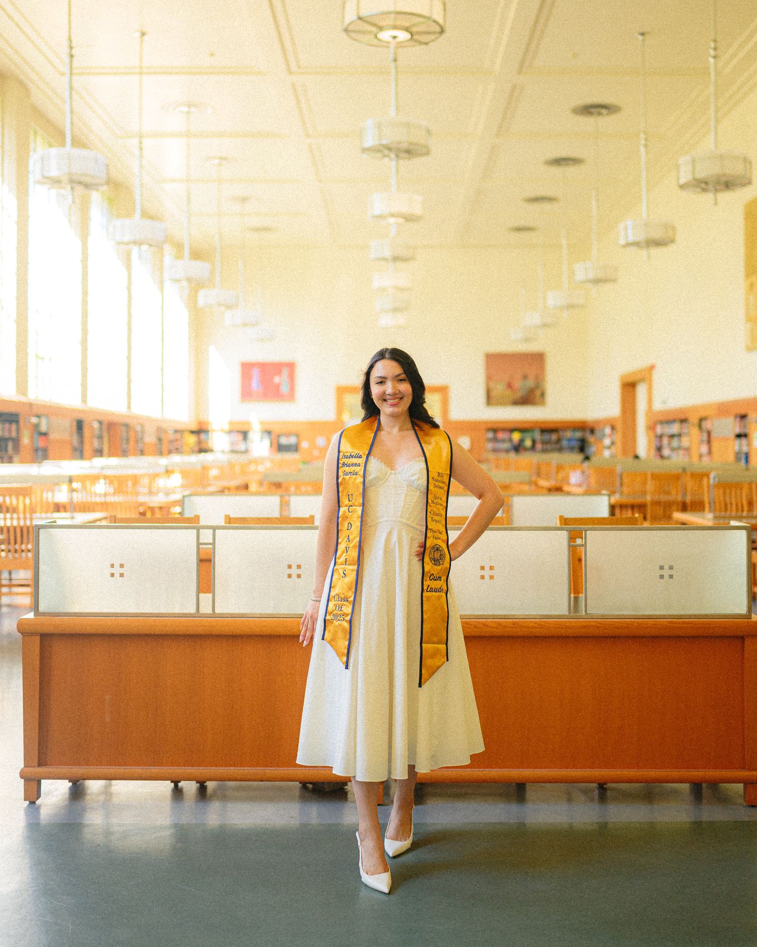 Graduate wearing white dress and gold stole stands in university library for graduation photo.