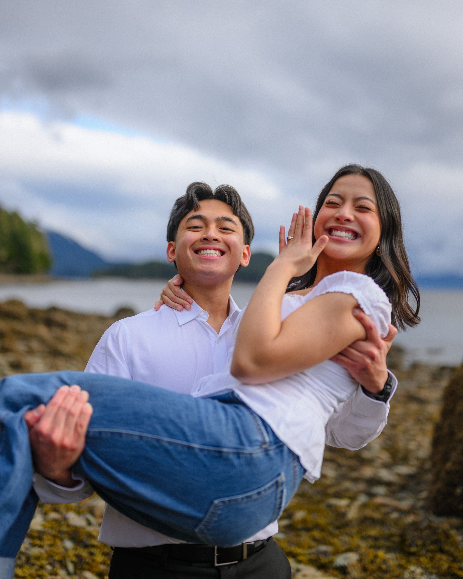 Couple in casual attire poses playfully on rocky coastal landscape.