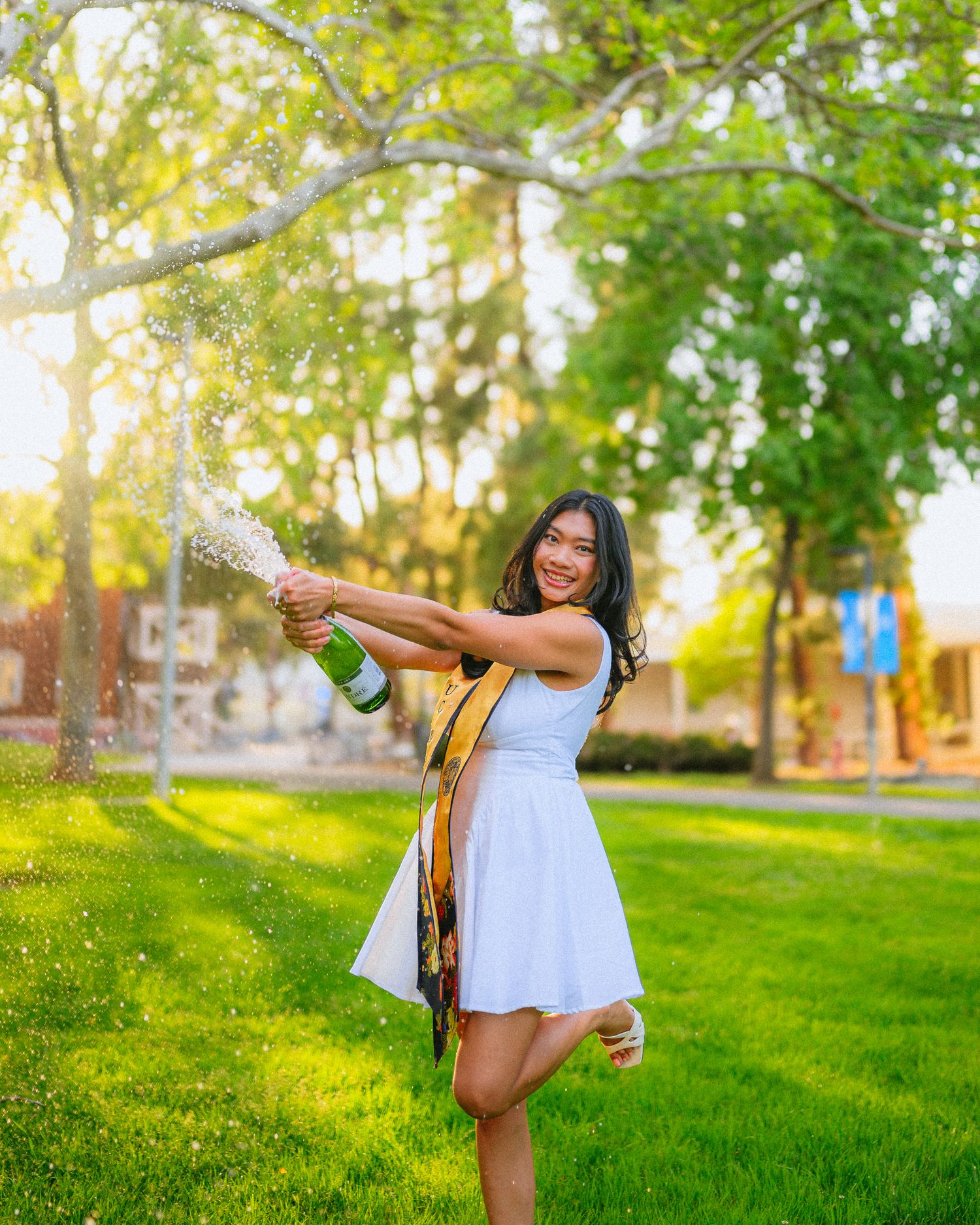 Joyful graduation photoshoot in a sunlit park with white dress and gold stole.